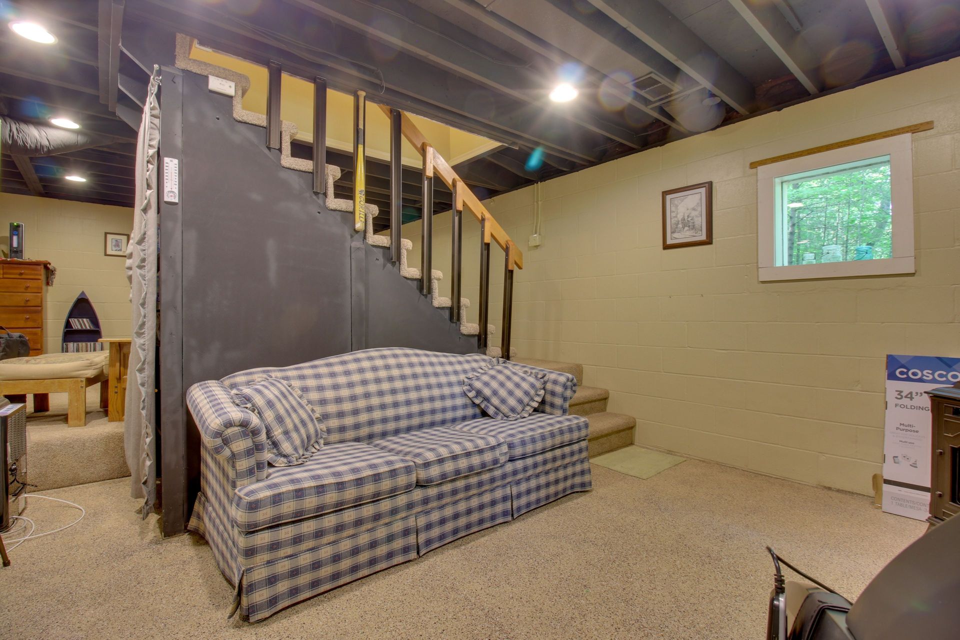 Basement with plaid couch under wooden staircase, small window on right.