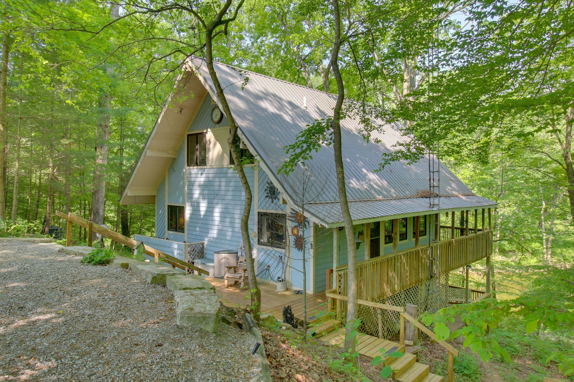 Blue A-frame cabin in a wooded setting with a wraparound porch and a gravel driveway.