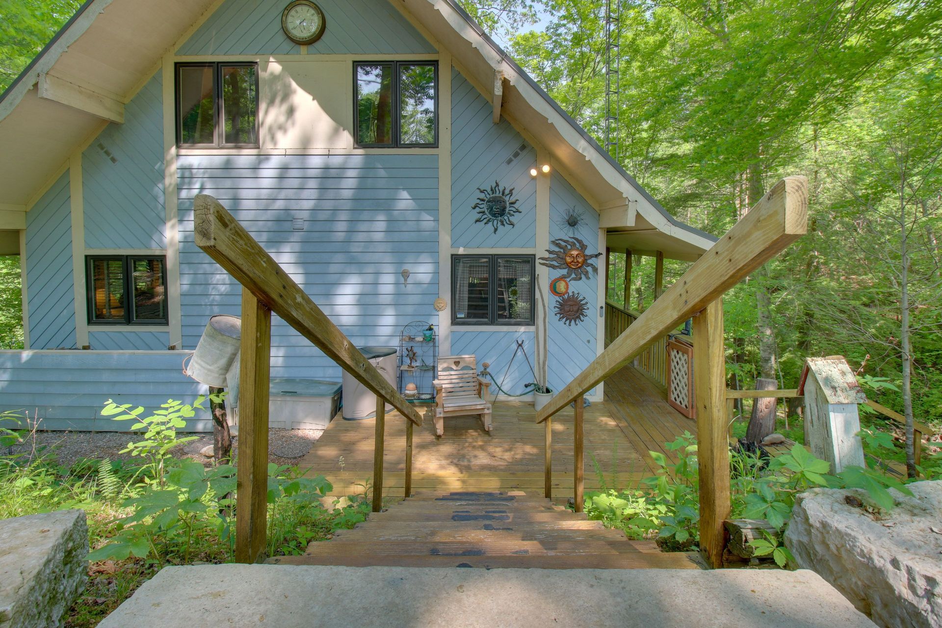 A-frame cabin, light blue siding, wooden stairs lead to front porch, surrounded by trees.