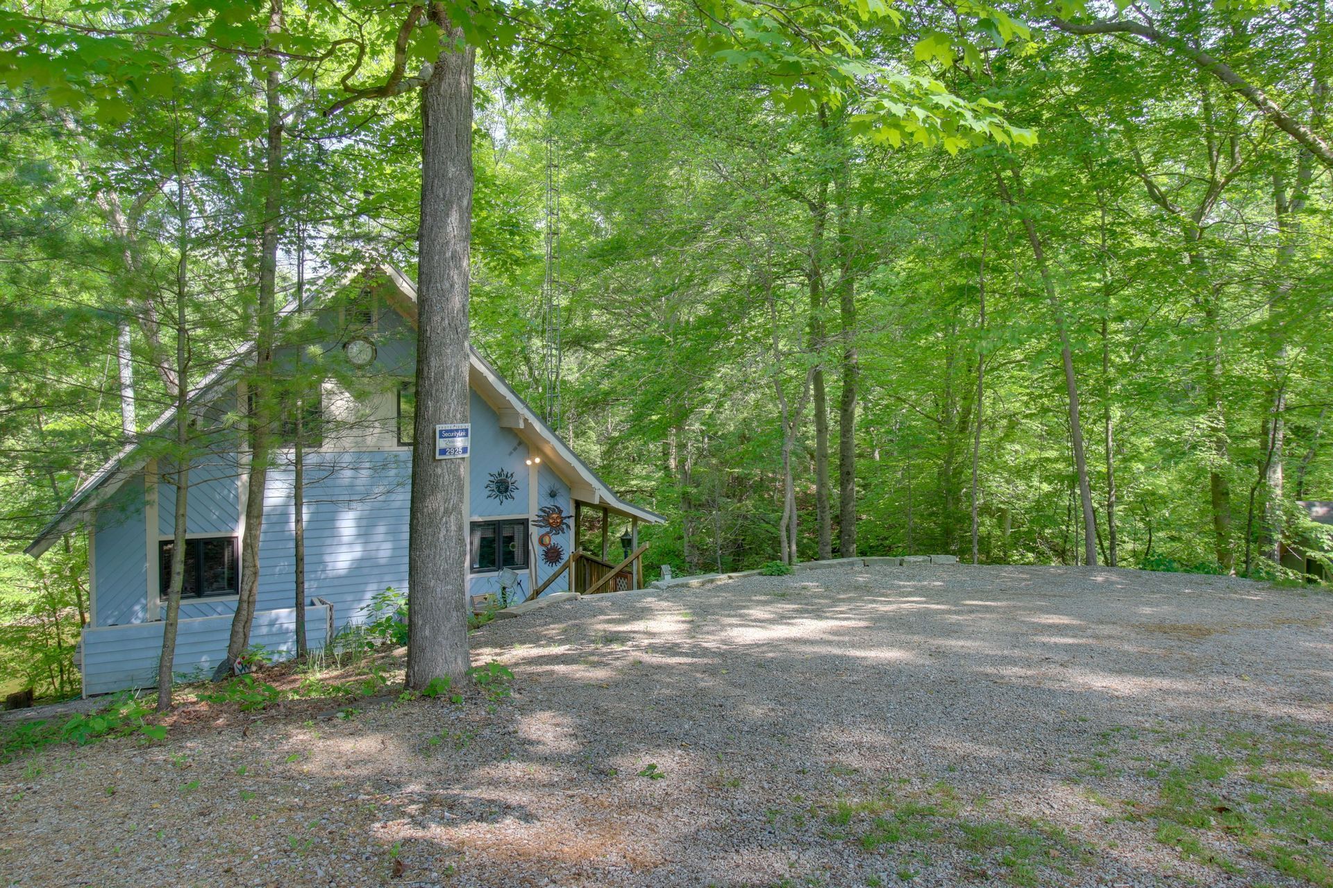 Blue cabin in a wooded area with gravel parking area.