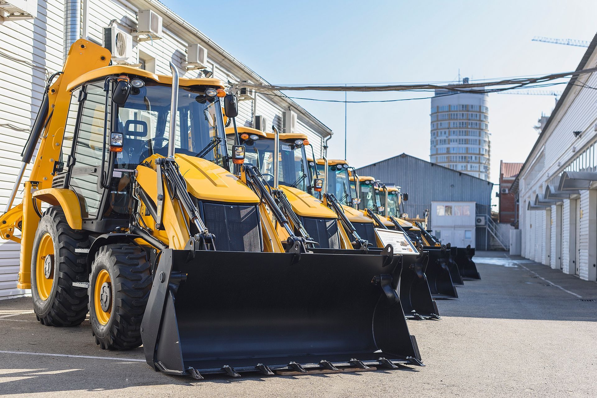Row of yellow backhoe loaders parked between industrial buildings.