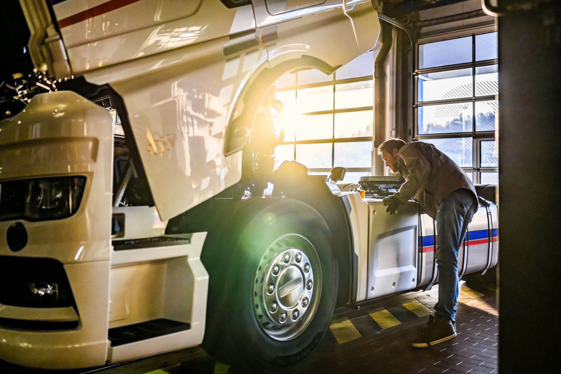 Mechanic inspecting a white semi-truck with hood open in a brightly lit garage.