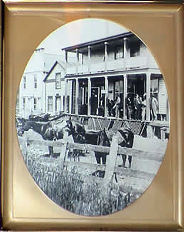 Oval framed vintage photo of a two-story building with a porch, people, and horses hitched to wagons in front.