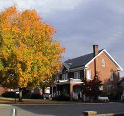 Brick house with a dark roof and porch. A large tree with yellow and orange leaves is in front. Cloudy sky.