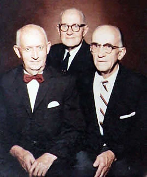 Three older men in suits posing for a portrait. Two have glasses, one with a bow tie.