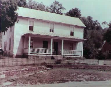 Two-story white house with a porch, metal fence, and trees in the background.