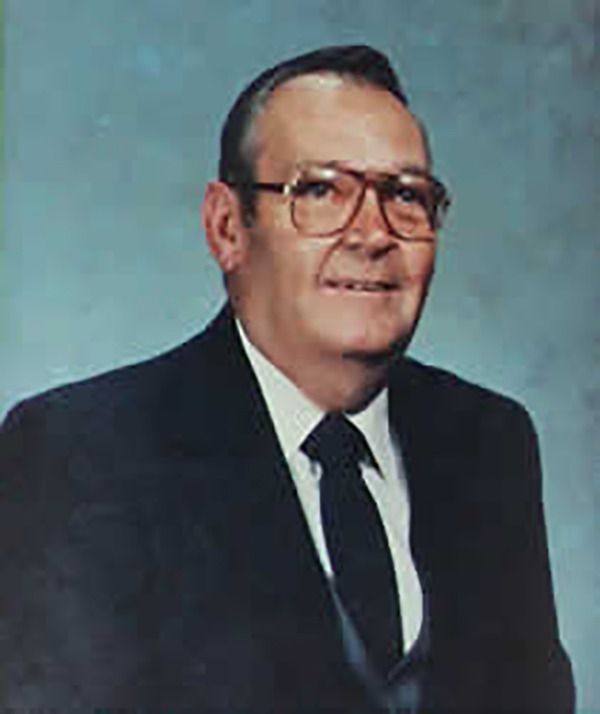 Man wearing glasses, a dark suit, and a tie, smiling against a light blue background.