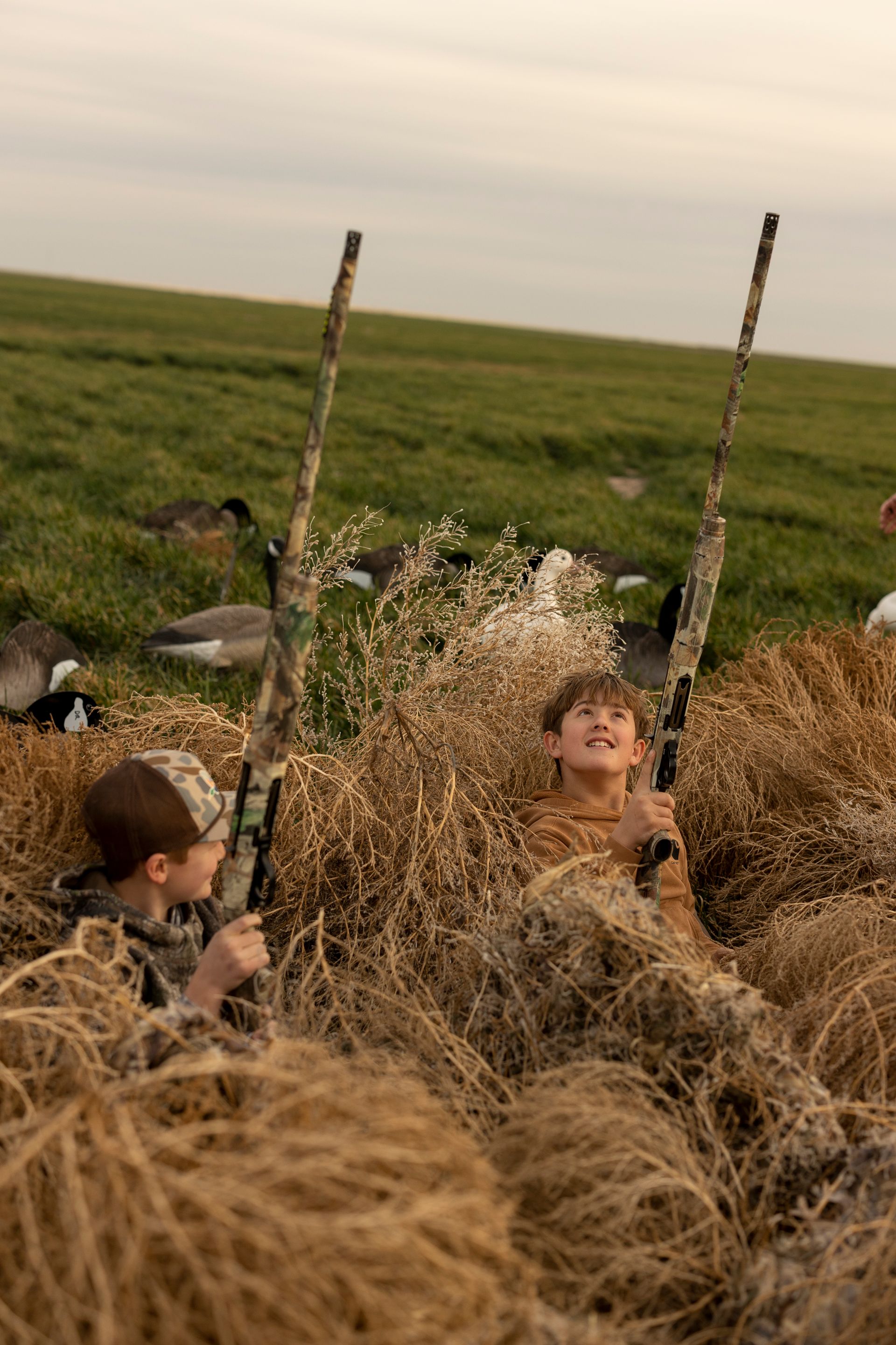 Two children concealed in tall grass, holding camouflage rifles, looking up at the sky.