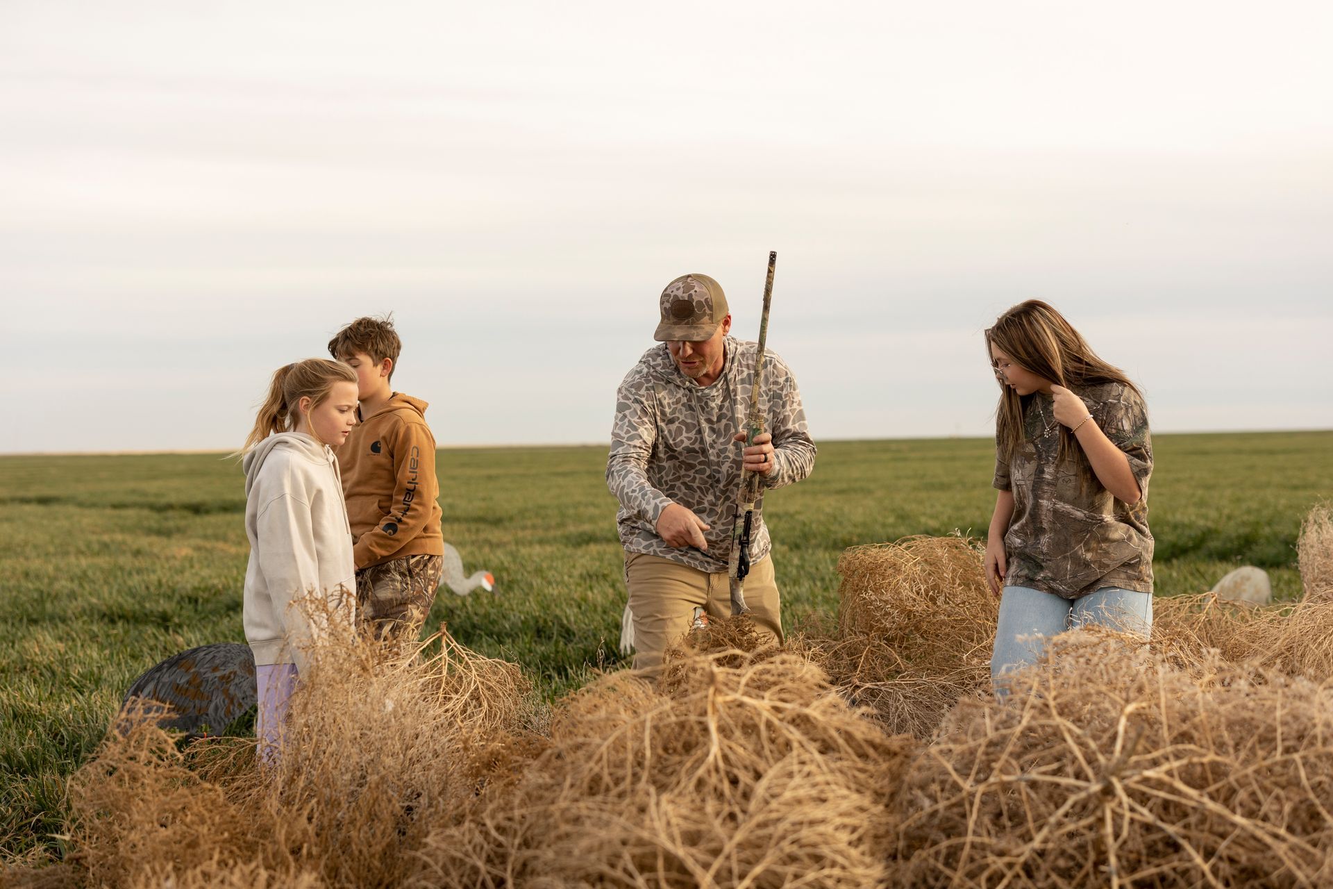 Family examining tumbleweeds in a field. Man in camo points; children look on. Overcast day.