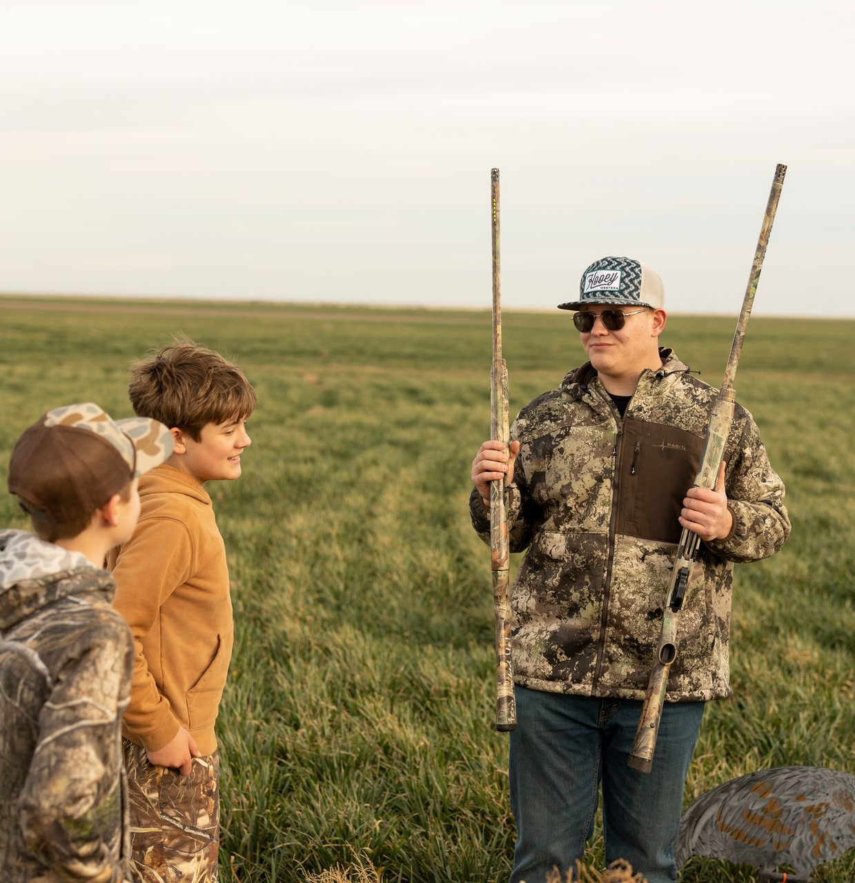 Person with two shotguns instructs two children in a field.