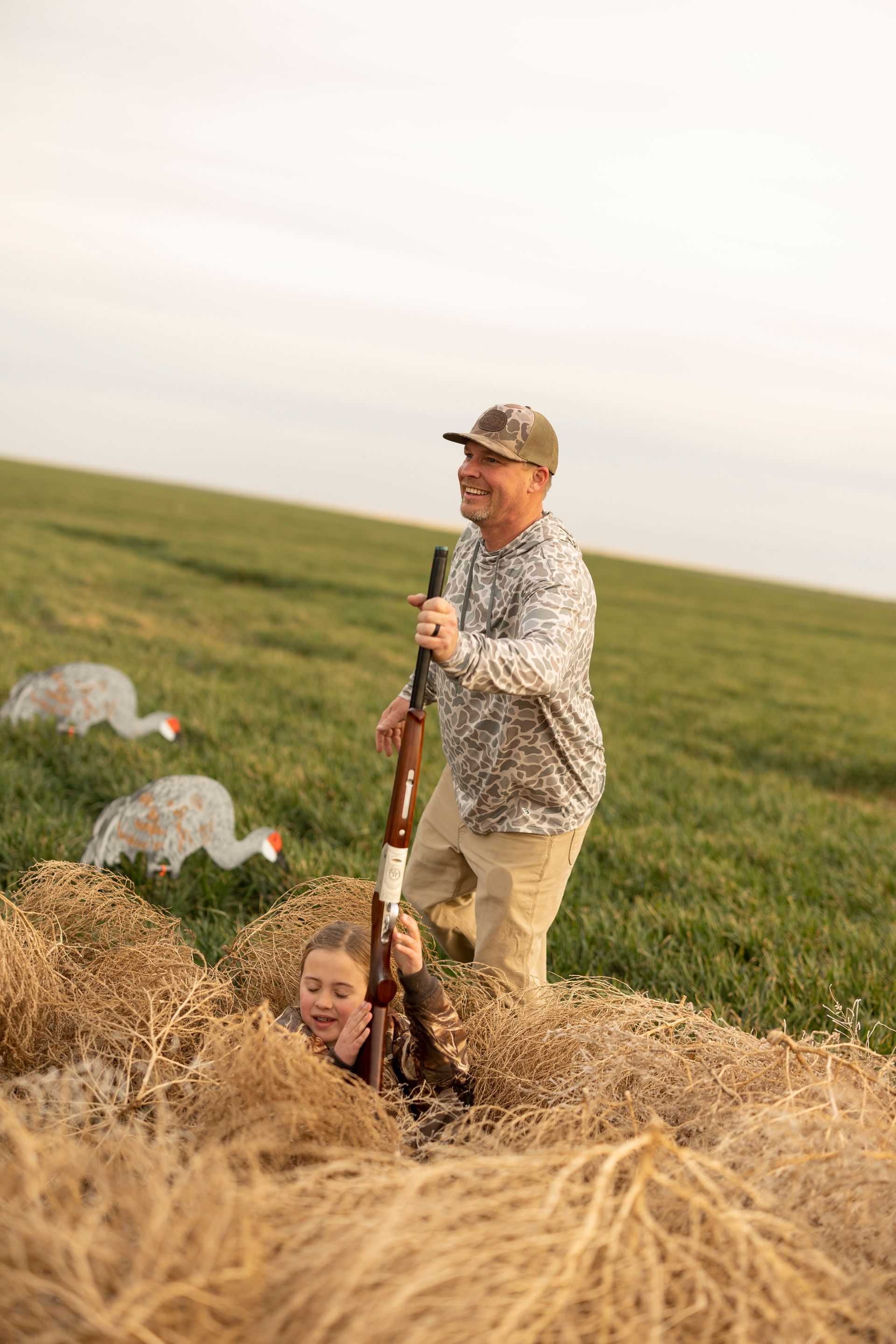 Man and child in hunting blind, holding a shotgun, smiling. Green field, decoys in background.