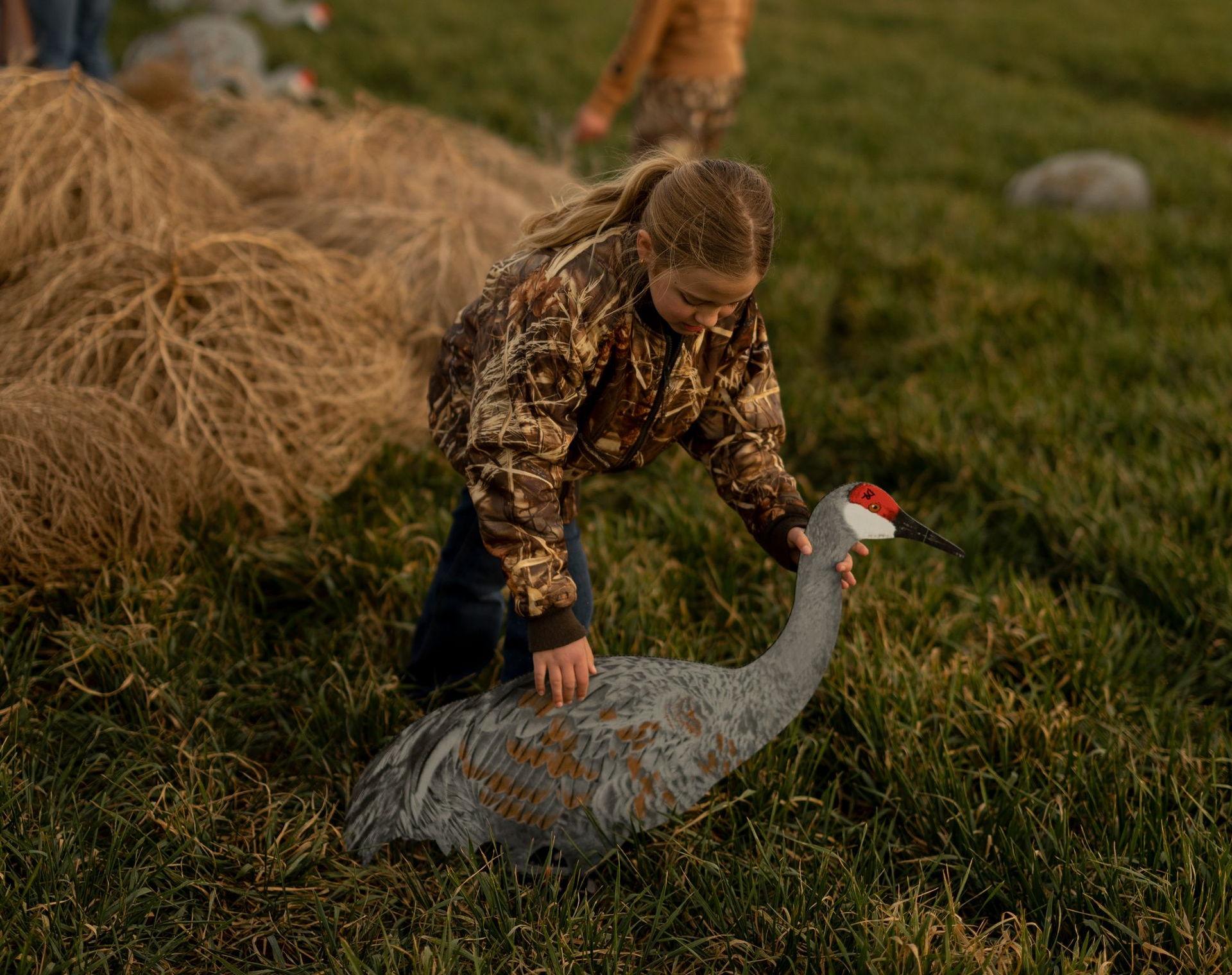 Girl in camo jacket touching a Sandhill crane decoy in a grassy field.