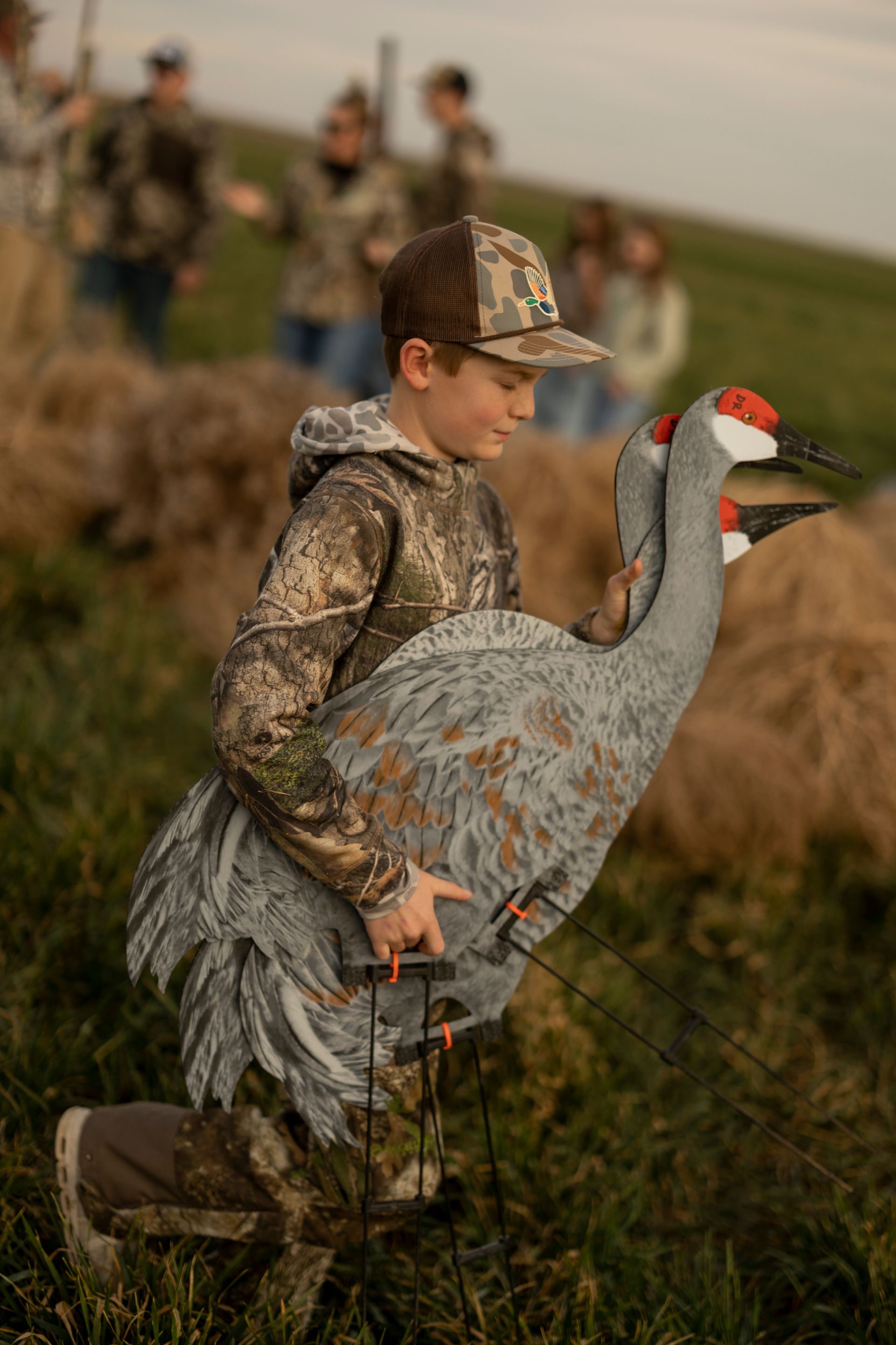 Boy kneels, carrying two crane decoys in a grassy field. Others in camouflage are in the background.