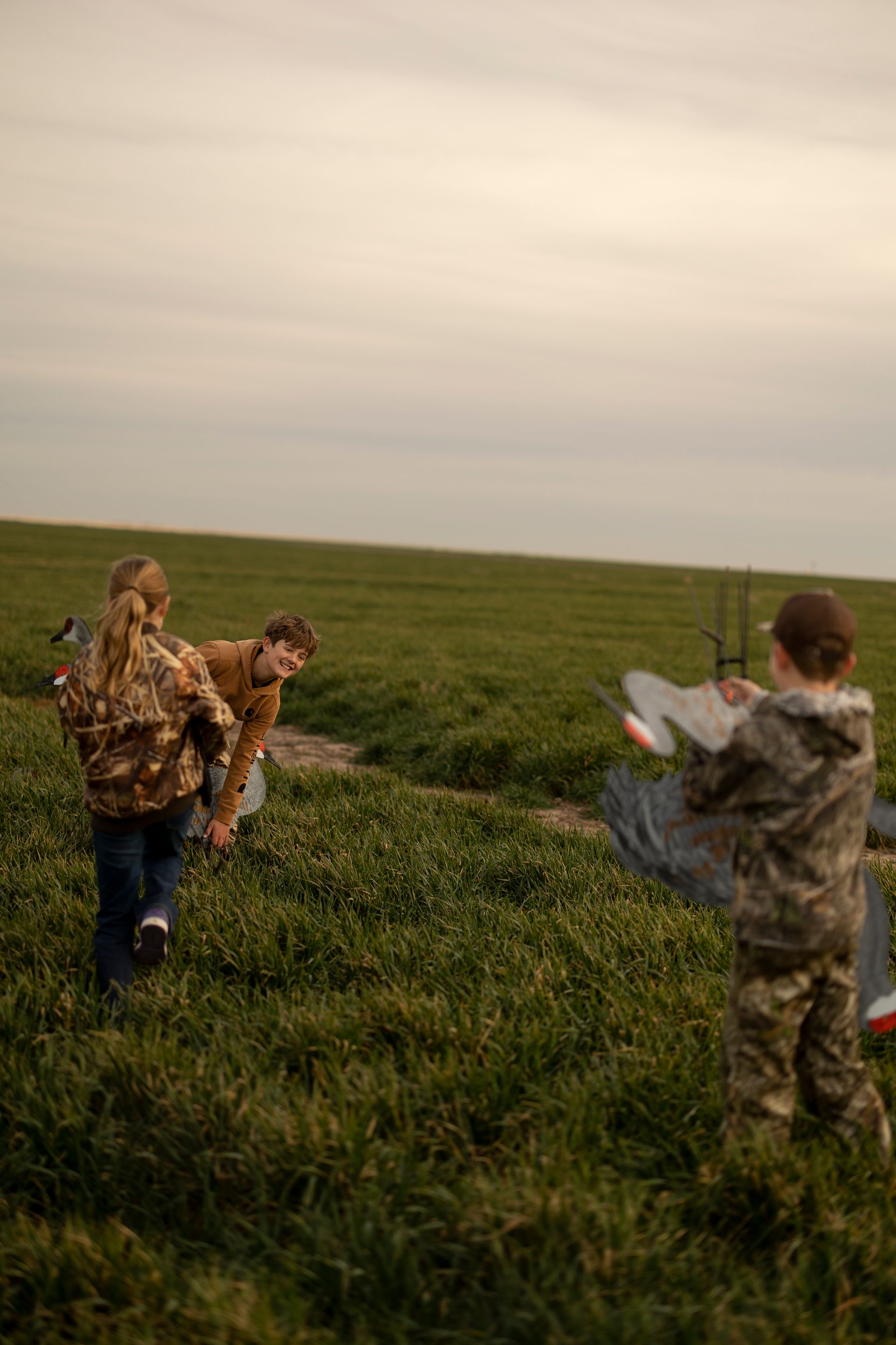 Children playing outdoors on a grassy field; one holds a gray object, others watch nearby. Overcast sky.