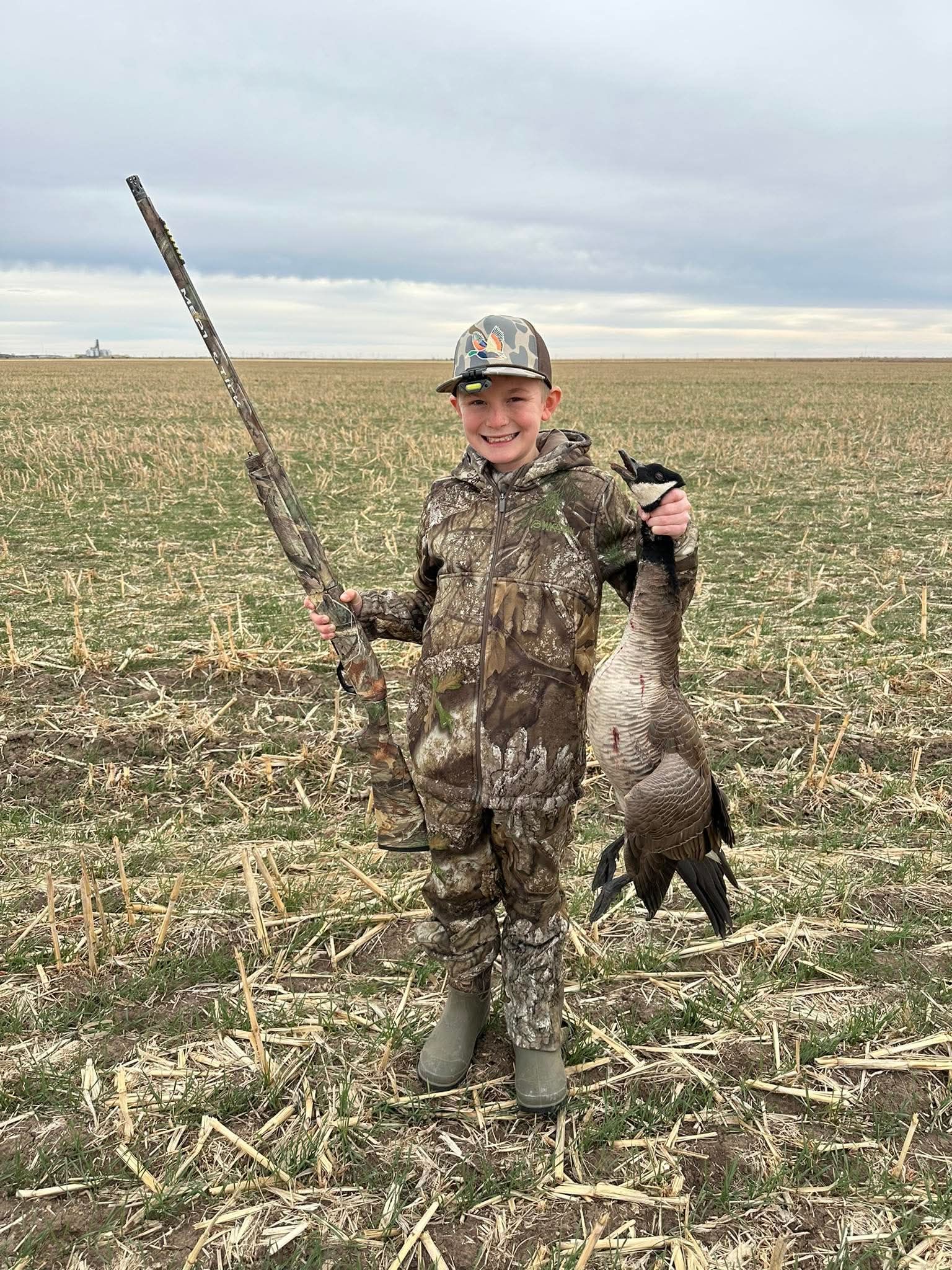 Young person in camouflage holds a shotgun and dead goose in a field.