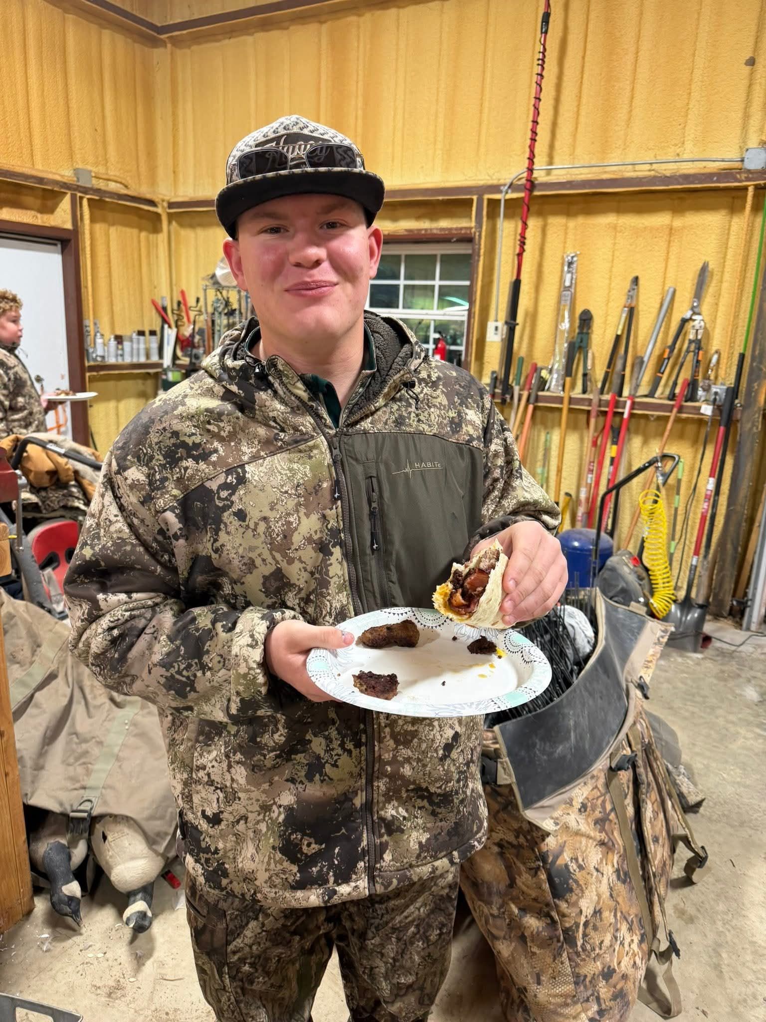 Person in camouflage eating a taco, smiling, indoors with hunting gear.