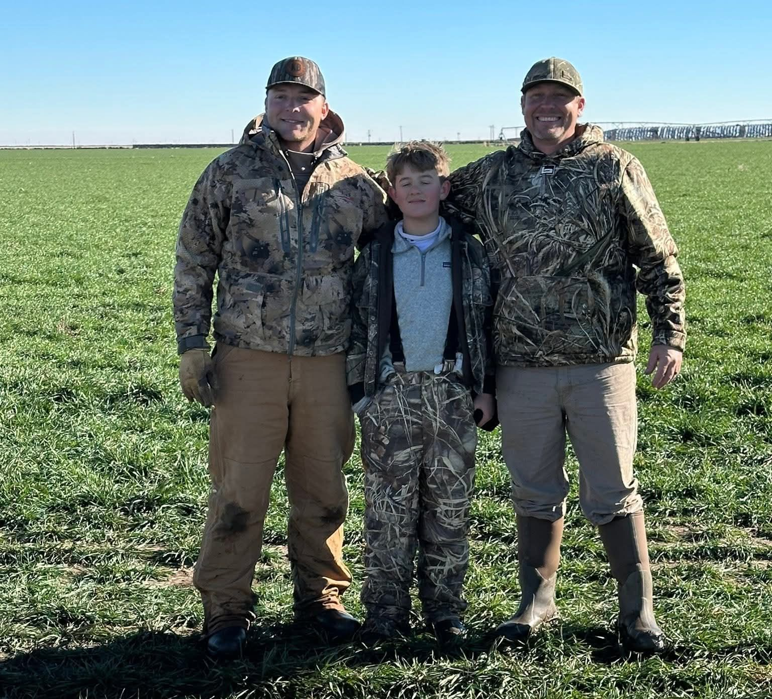 Three people in camouflage gear stand in a field of green plants under a blue sky.