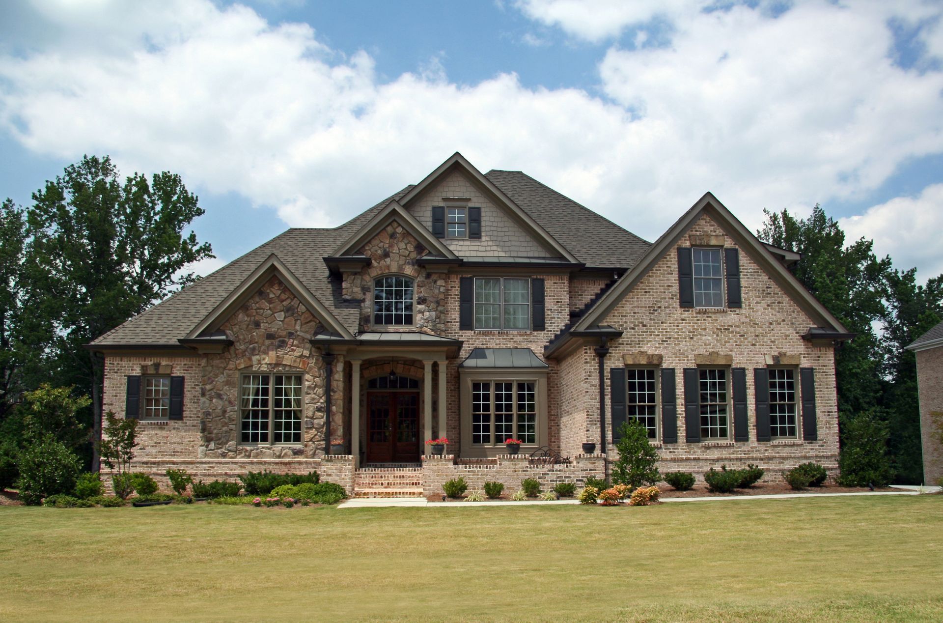 Two-story brick house with dark shutters, front porch, and grassy lawn under a partly cloudy sky.