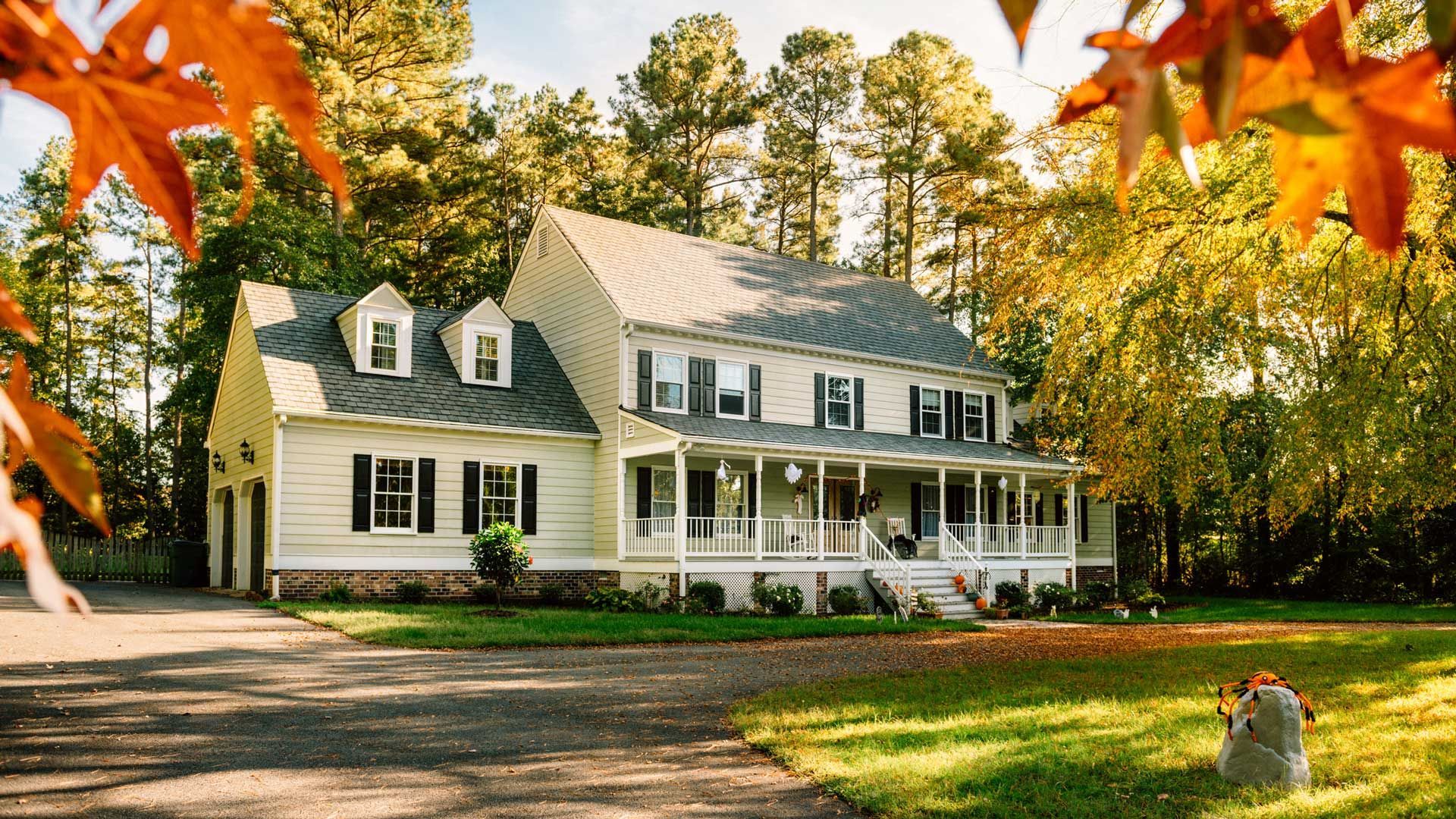 Two-story white house with a porch and black shutters in a fall setting, surrounded by trees with yellow and orange leaves.