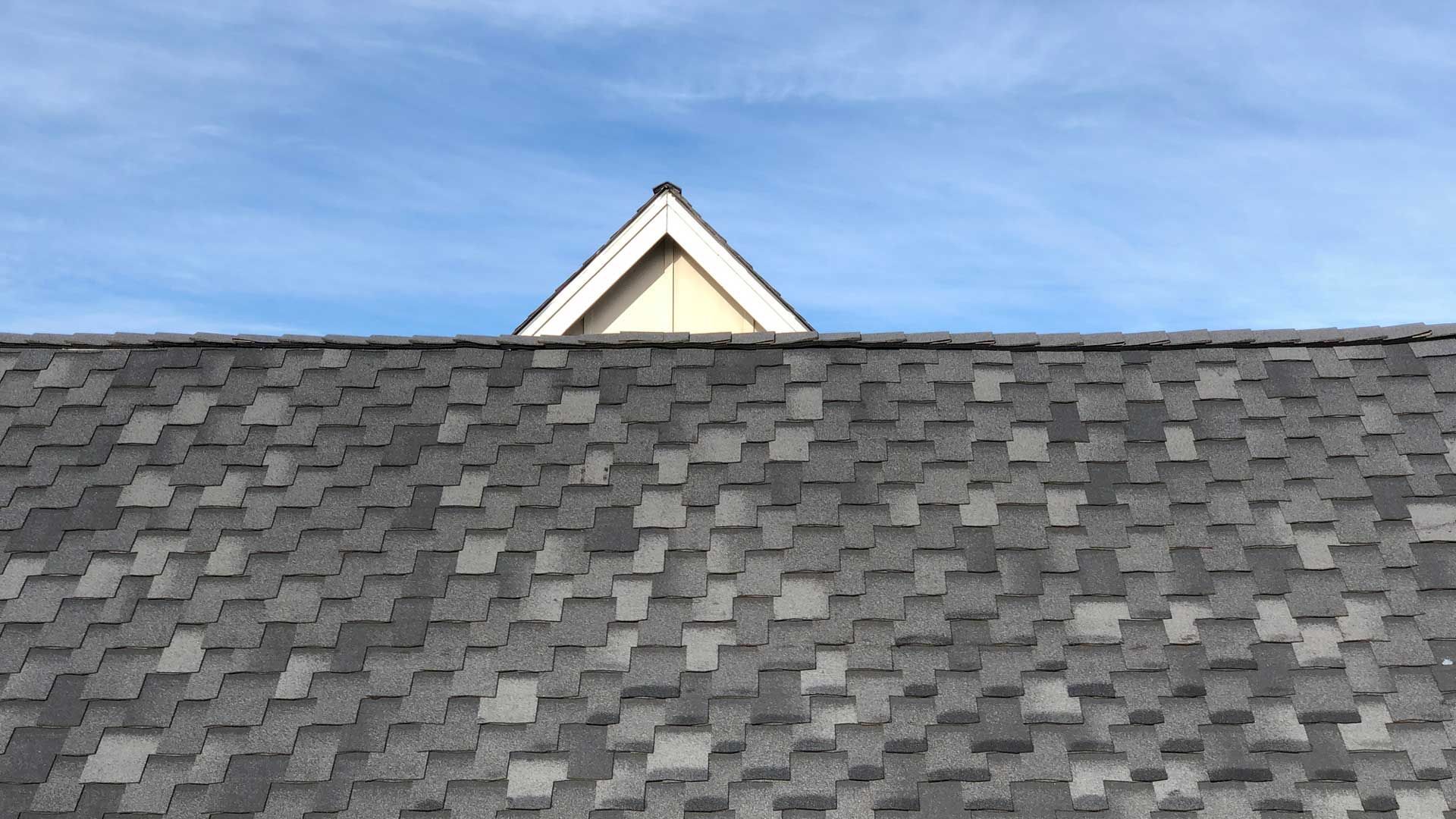 Gray asphalt shingle roof with a triangular roof peaking over it against a blue sky.