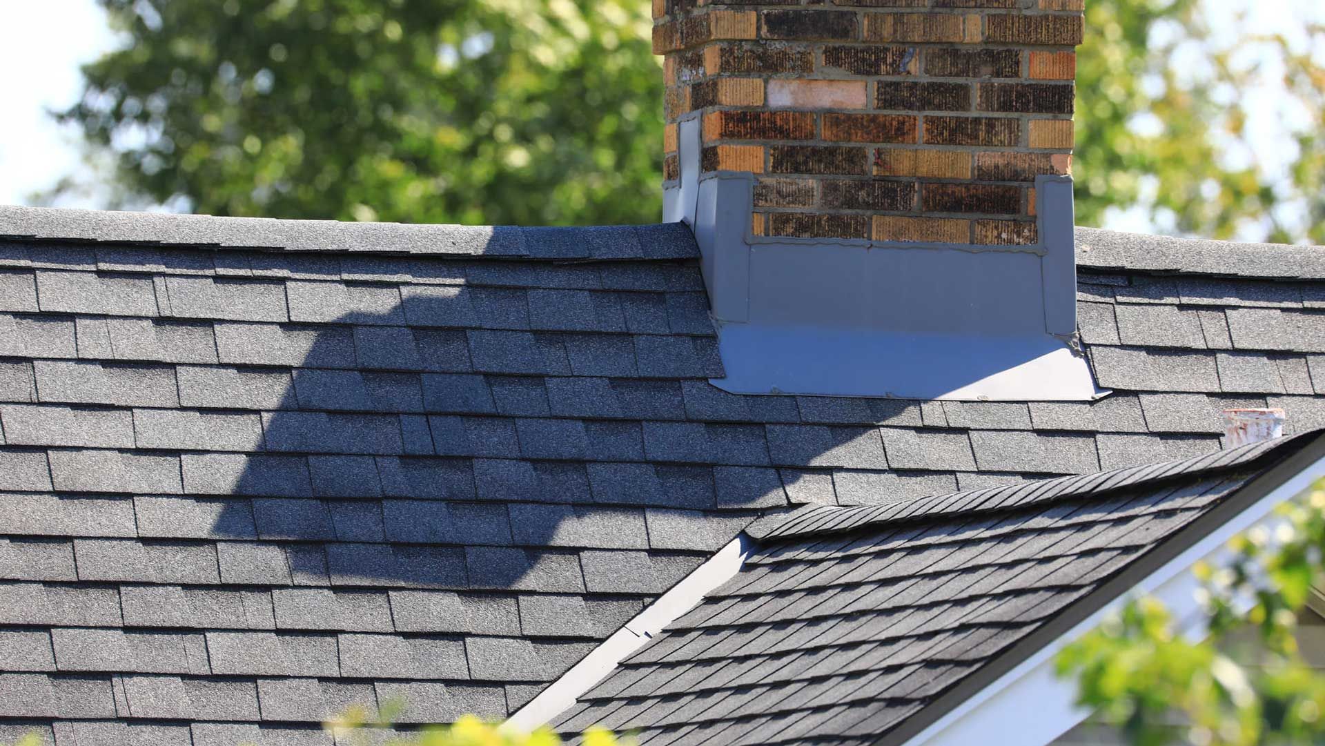 Dark gray shingled roof with a brick chimney and gray metal flashing. Green trees in the background.