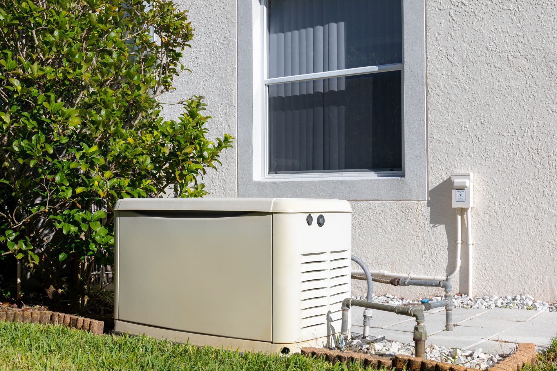 Beige home backup generator beside a house with a window and electrical outlet.