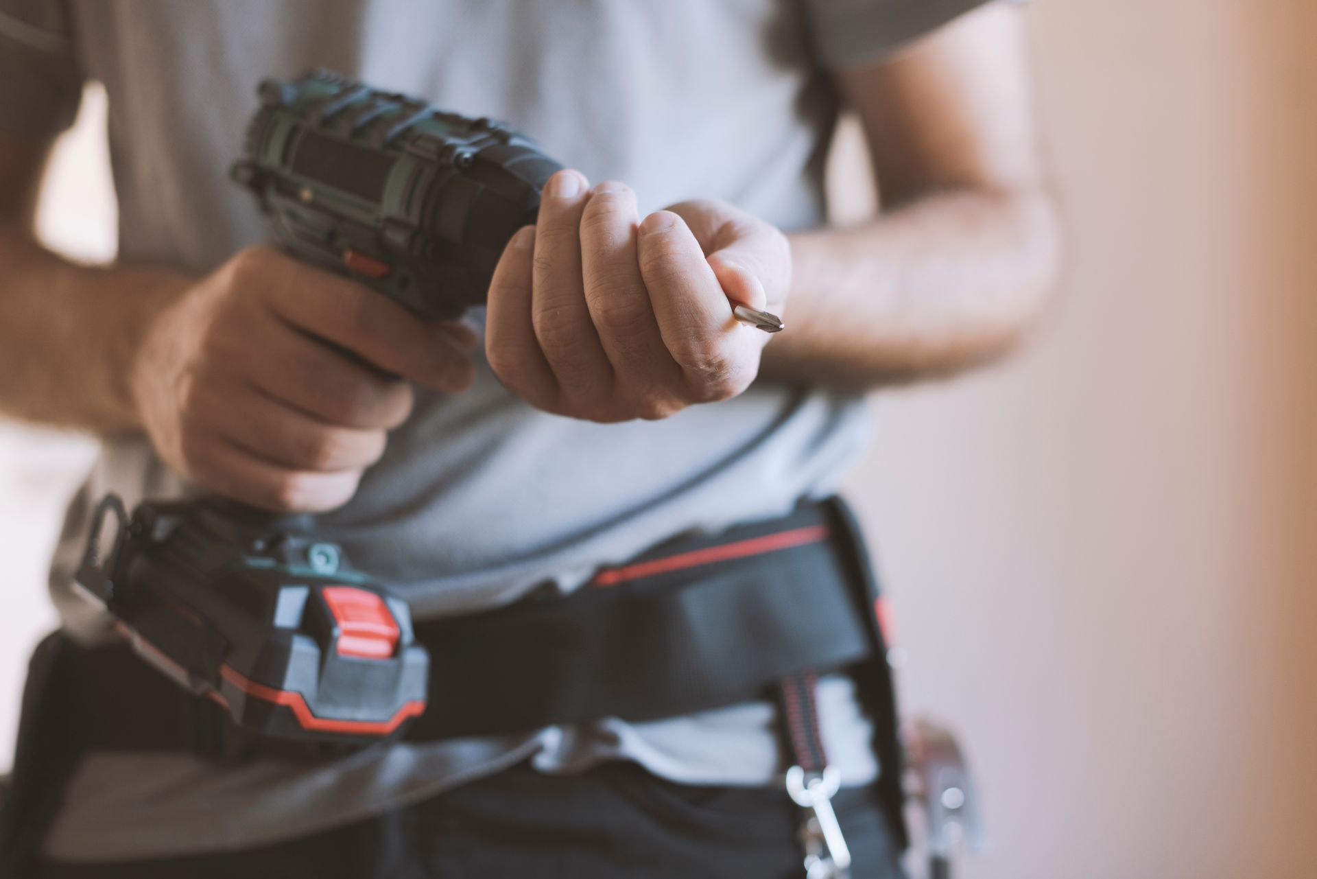 Person holding a screw, with a drill in one hand and tool belt, indoors.