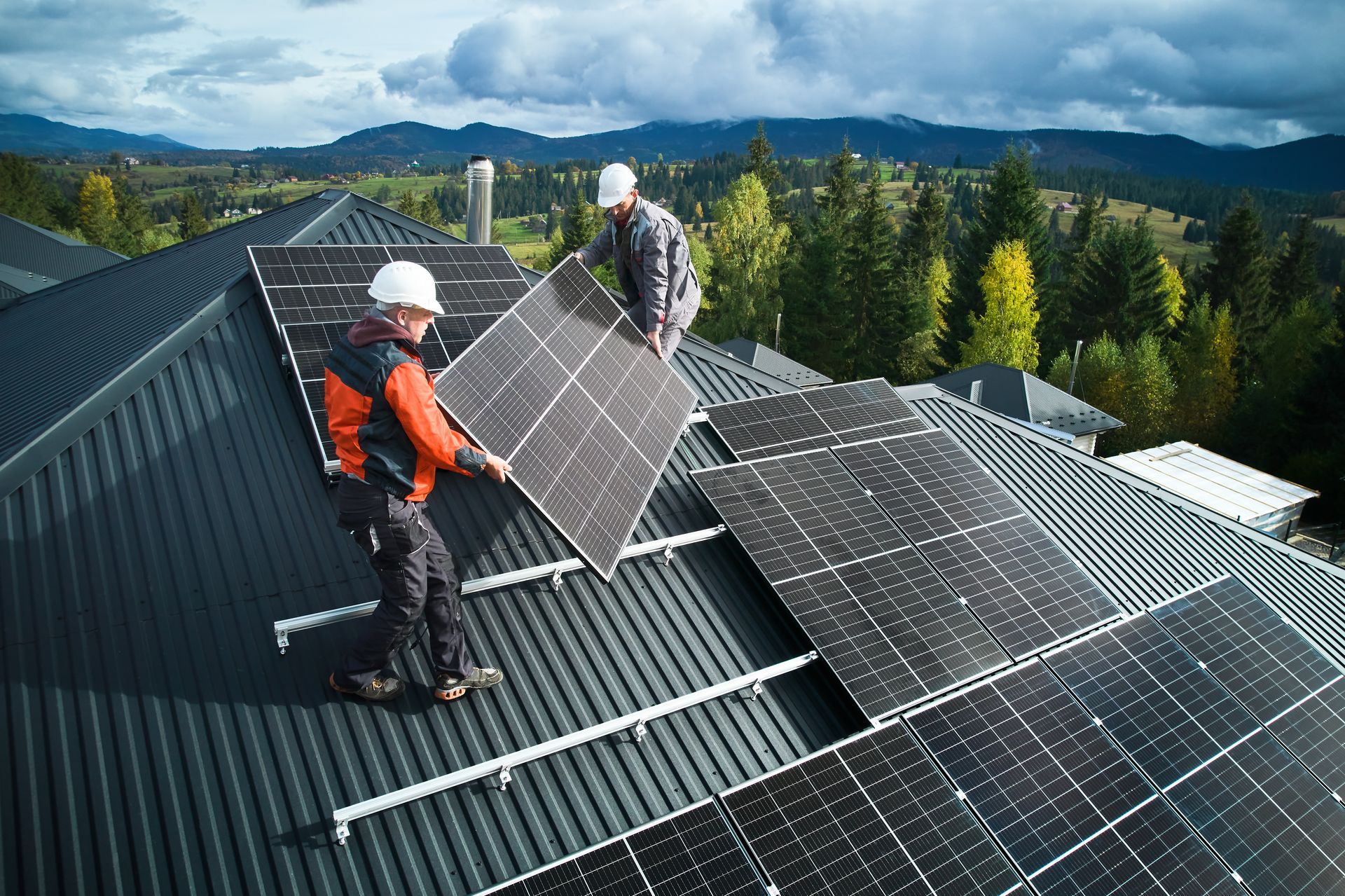 Two workers install solar panels on a rooftop. Mountains and trees are in the background.