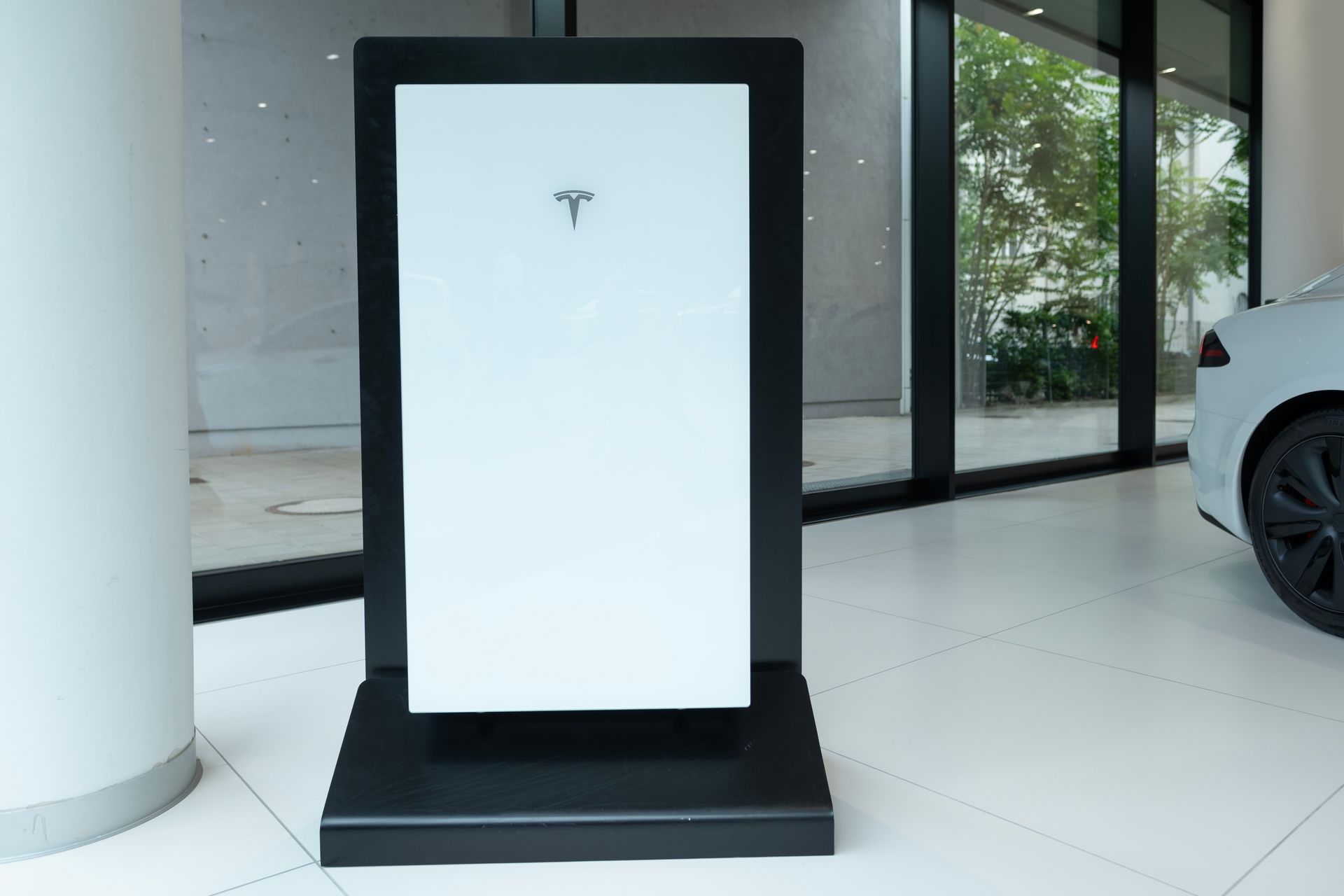 White Tesla Supercharger pedestal indoors near a glass entrance, with a car parked beside it