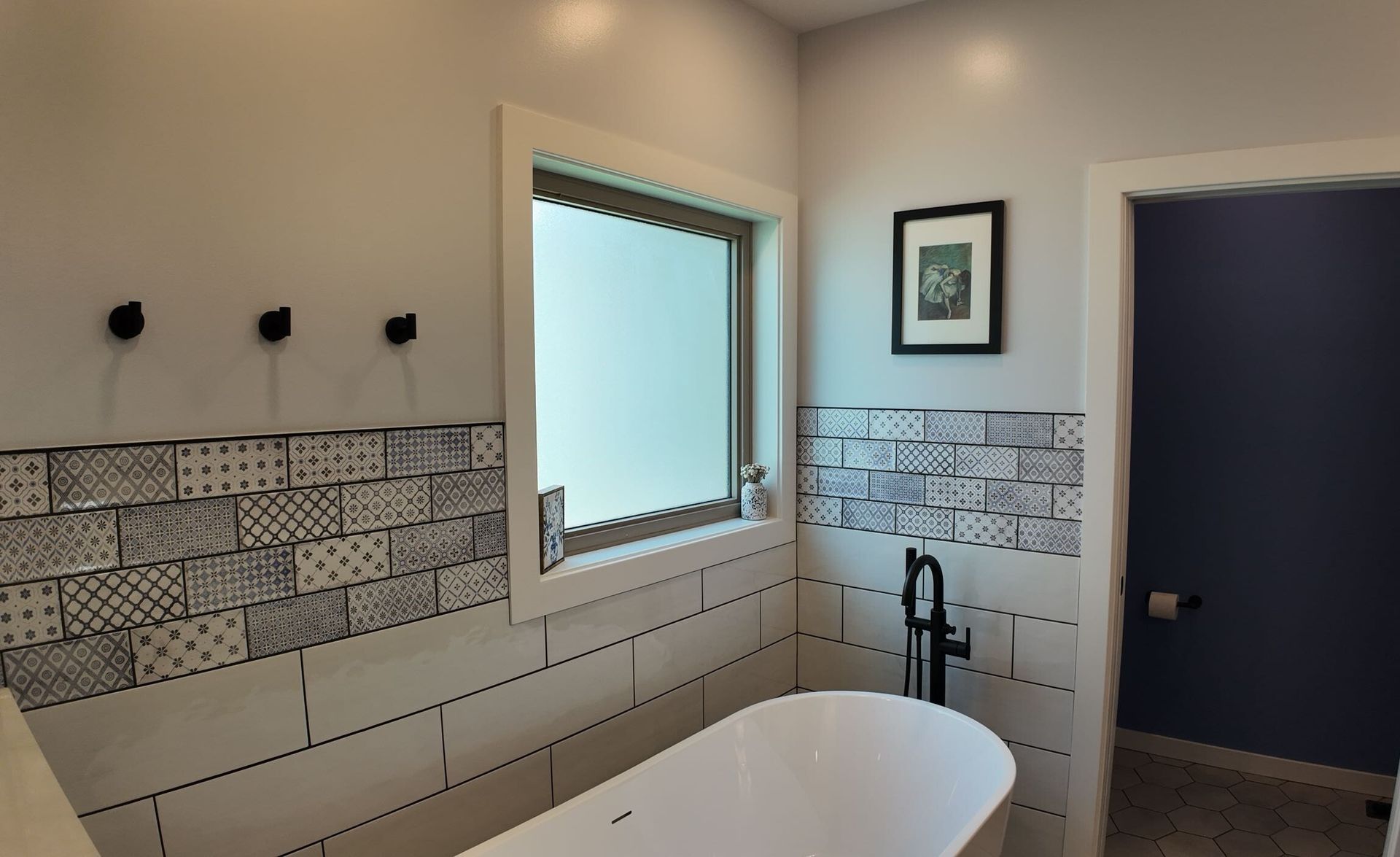 Bathroom with white tub, patterned tile, small window, black fixtures and a framed picture.