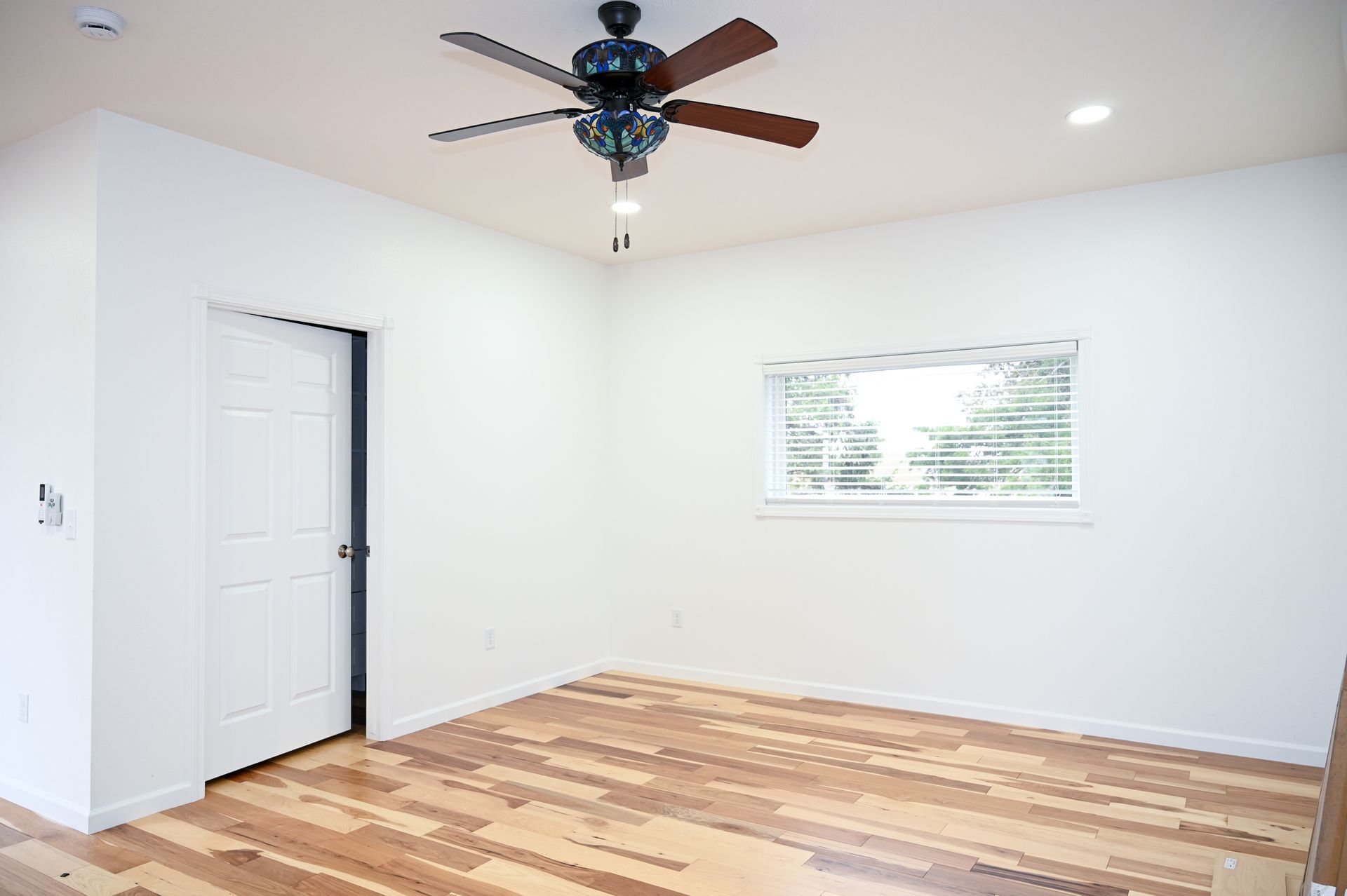 Empty room with wood floor, white walls, ceiling fan, window, and closed white door.