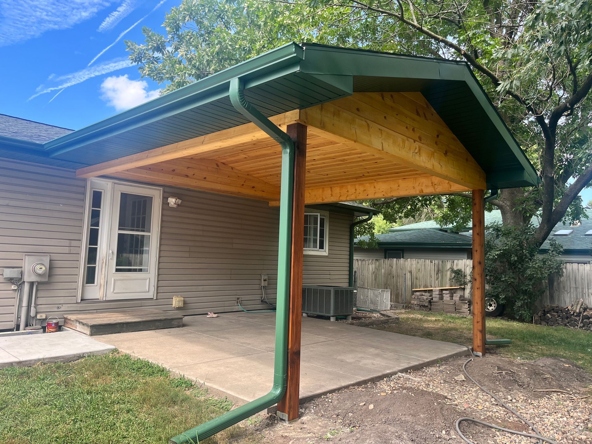 A covered patio with a wooden ceiling, green trim, and support posts. It's next to a house with a concrete slab.
