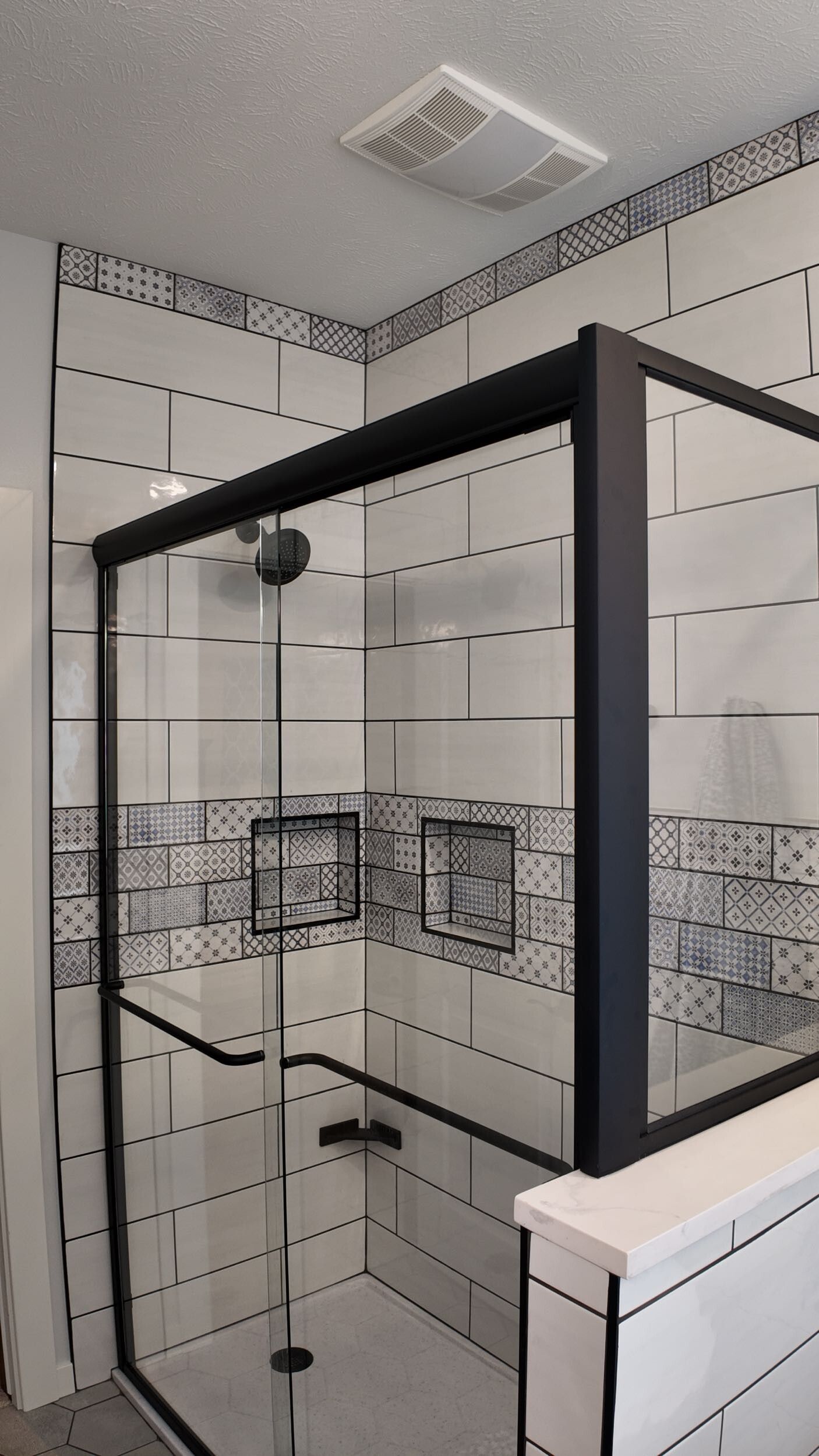 Black-framed glass shower enclosure with white tiled walls and a vent fan in a bathroom.