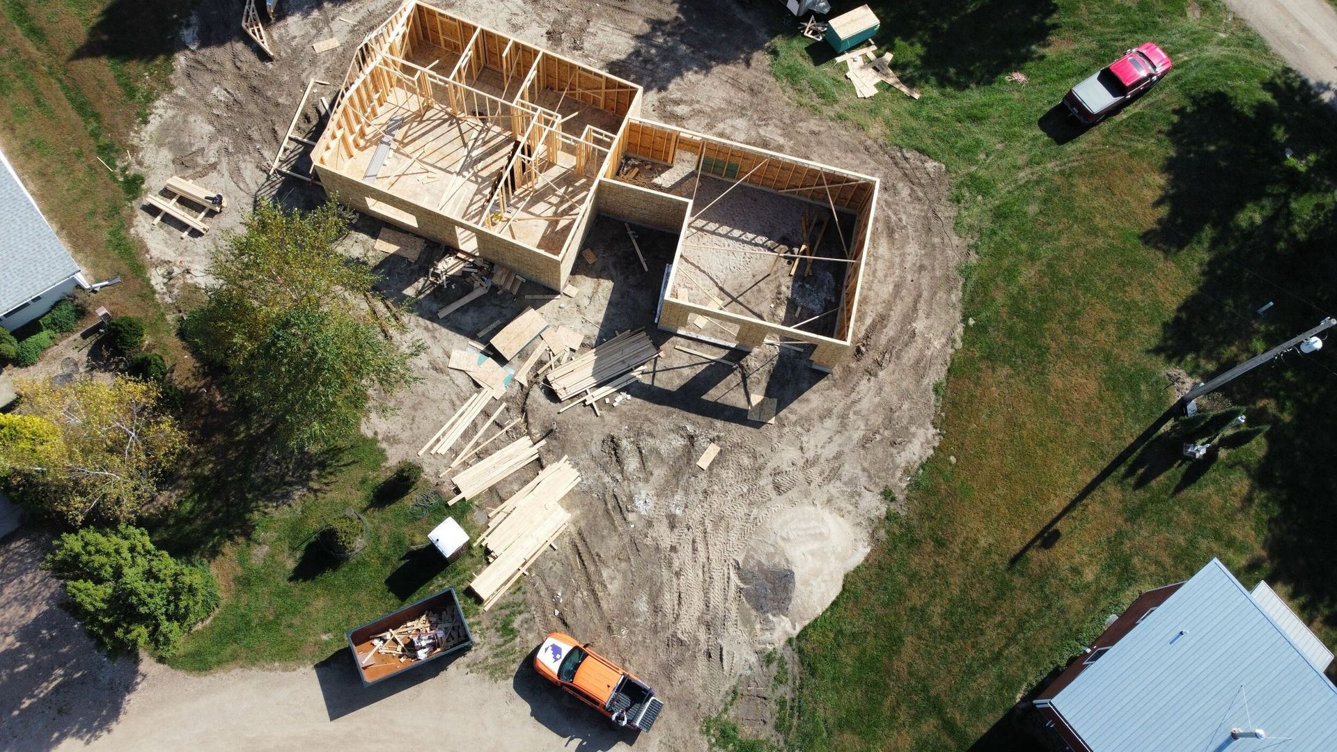 Aerial view of a home under construction; wooden frame on dirt lot, surrounded by construction materials, vehicles, and grass.