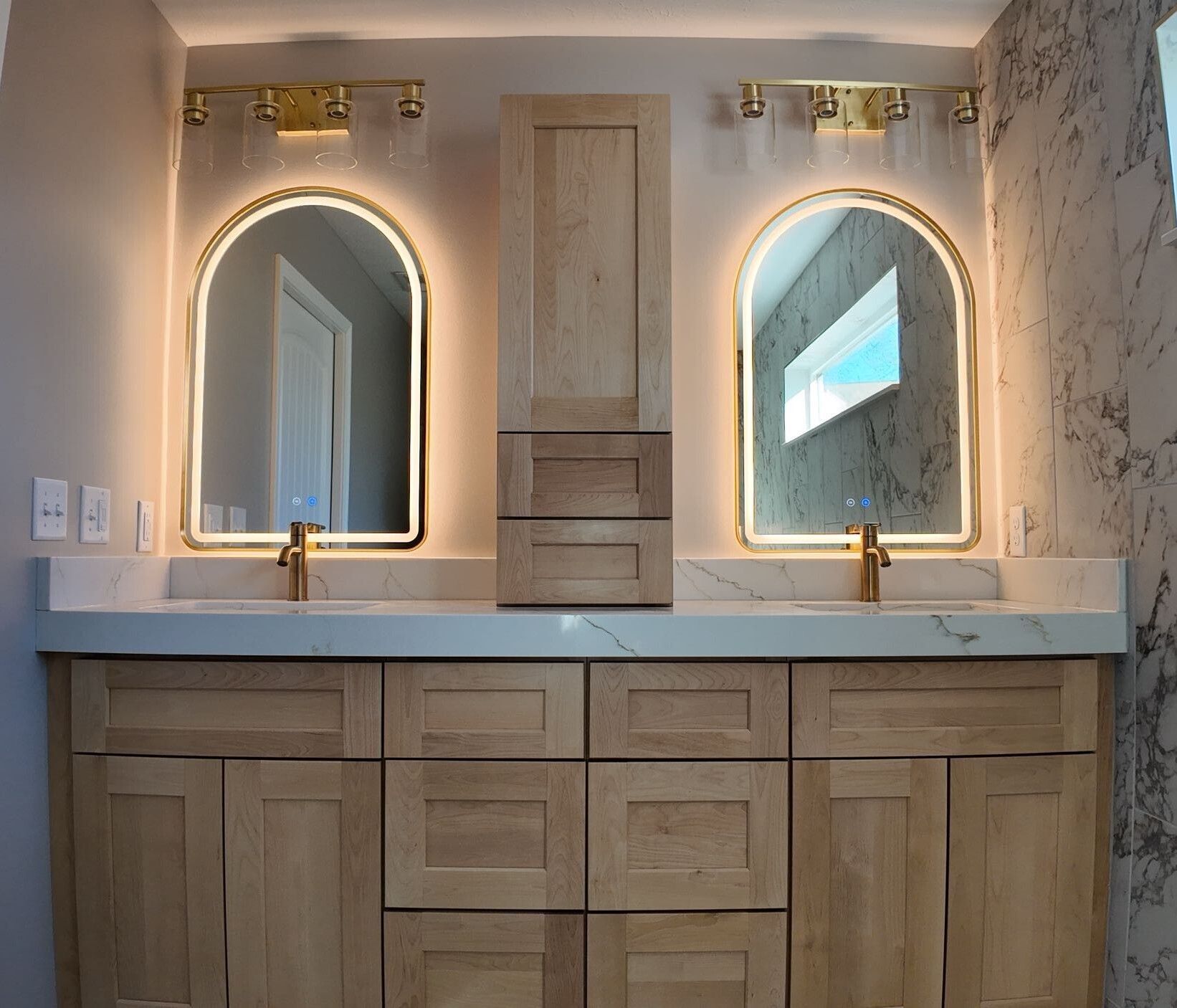 Bathroom vanity with two arched, lit mirrors, gold fixtures, and light wood cabinetry.