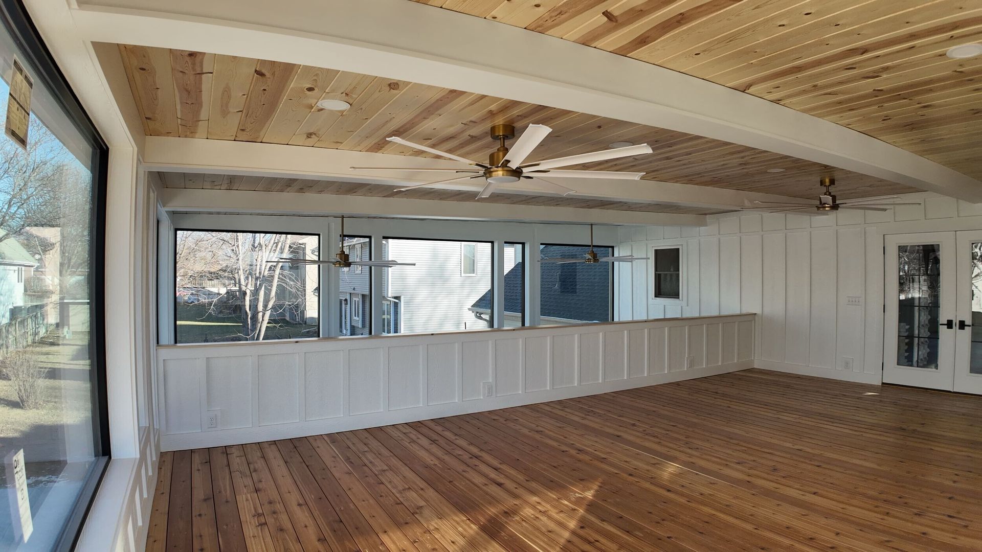 Sunroom interior with wood floors, paneled walls, and ceiling with fans and large windows.