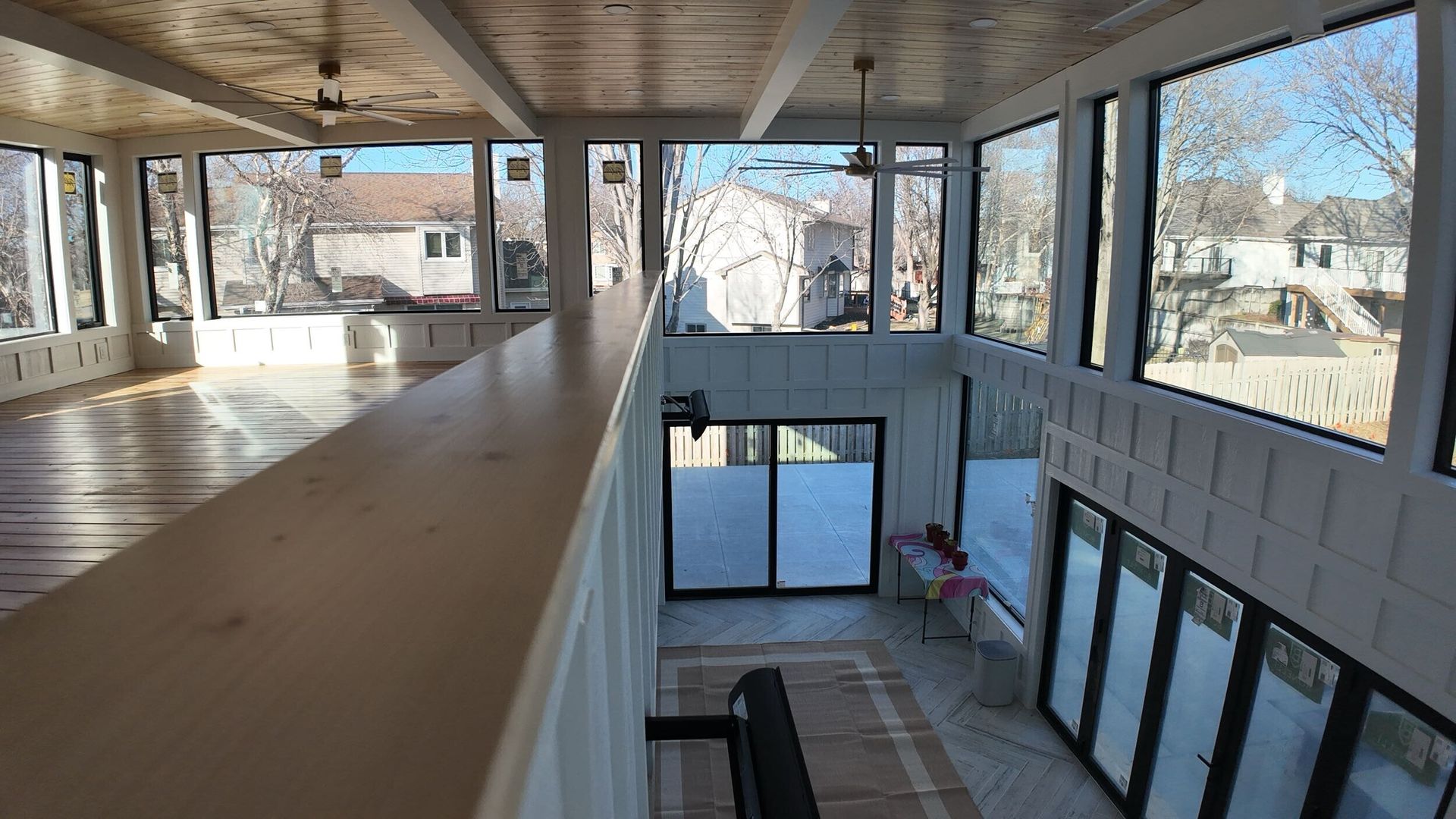 Interior view of a light-filled sunroom with large windows, wooden floors, and a balcony overlooking a lower level.