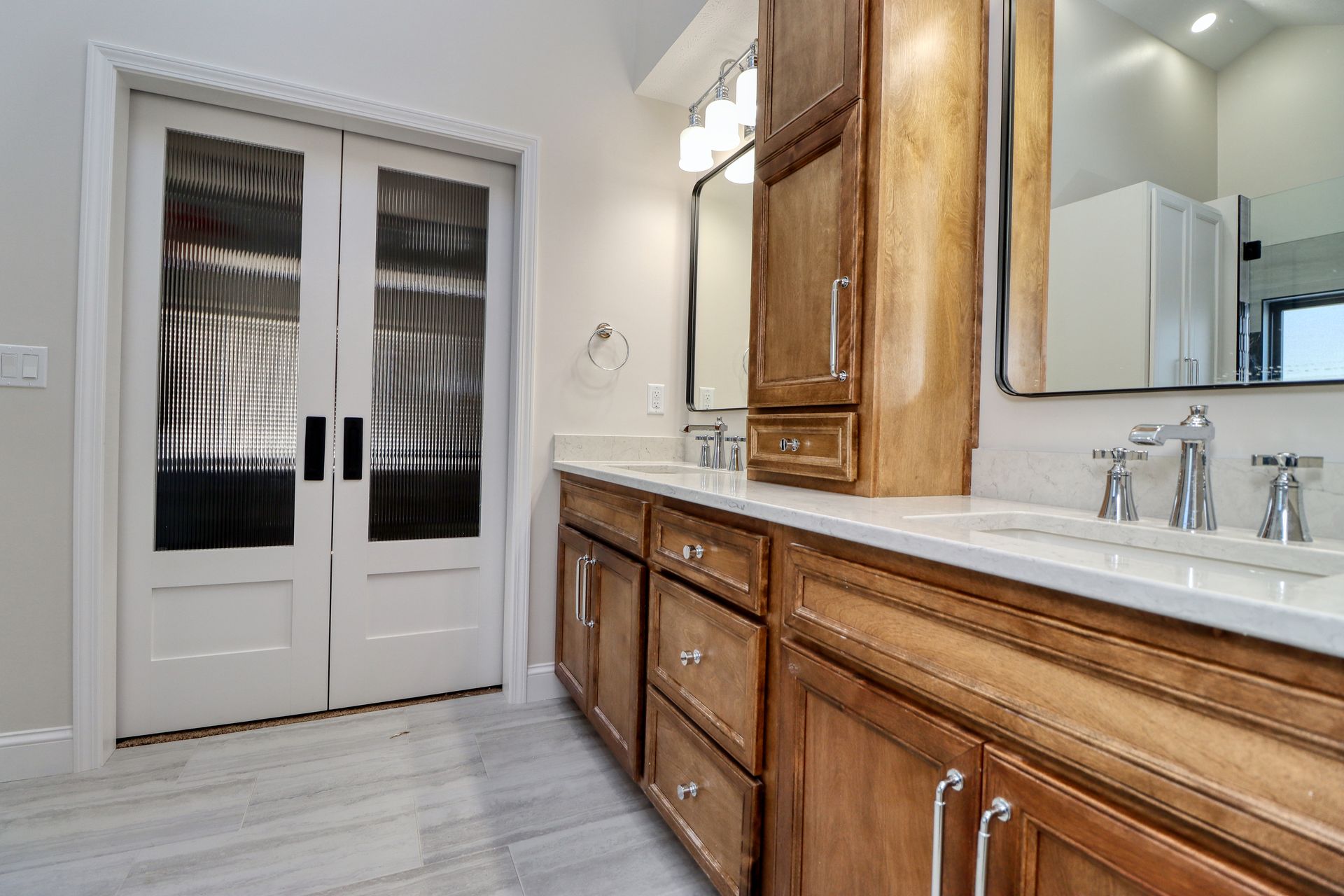 Bathroom with double doors, vanity with two sinks, and a tall cabinet. Light wood tones and white countertops.