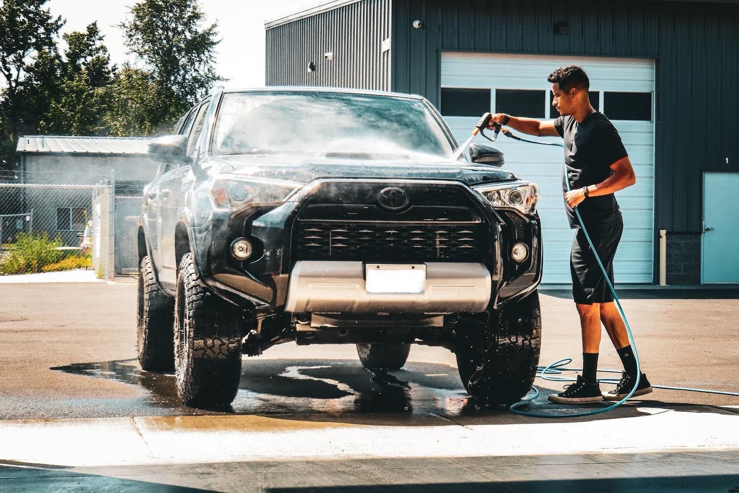 Man washes a black Toyota truck with water. Outdoors, sunlight.