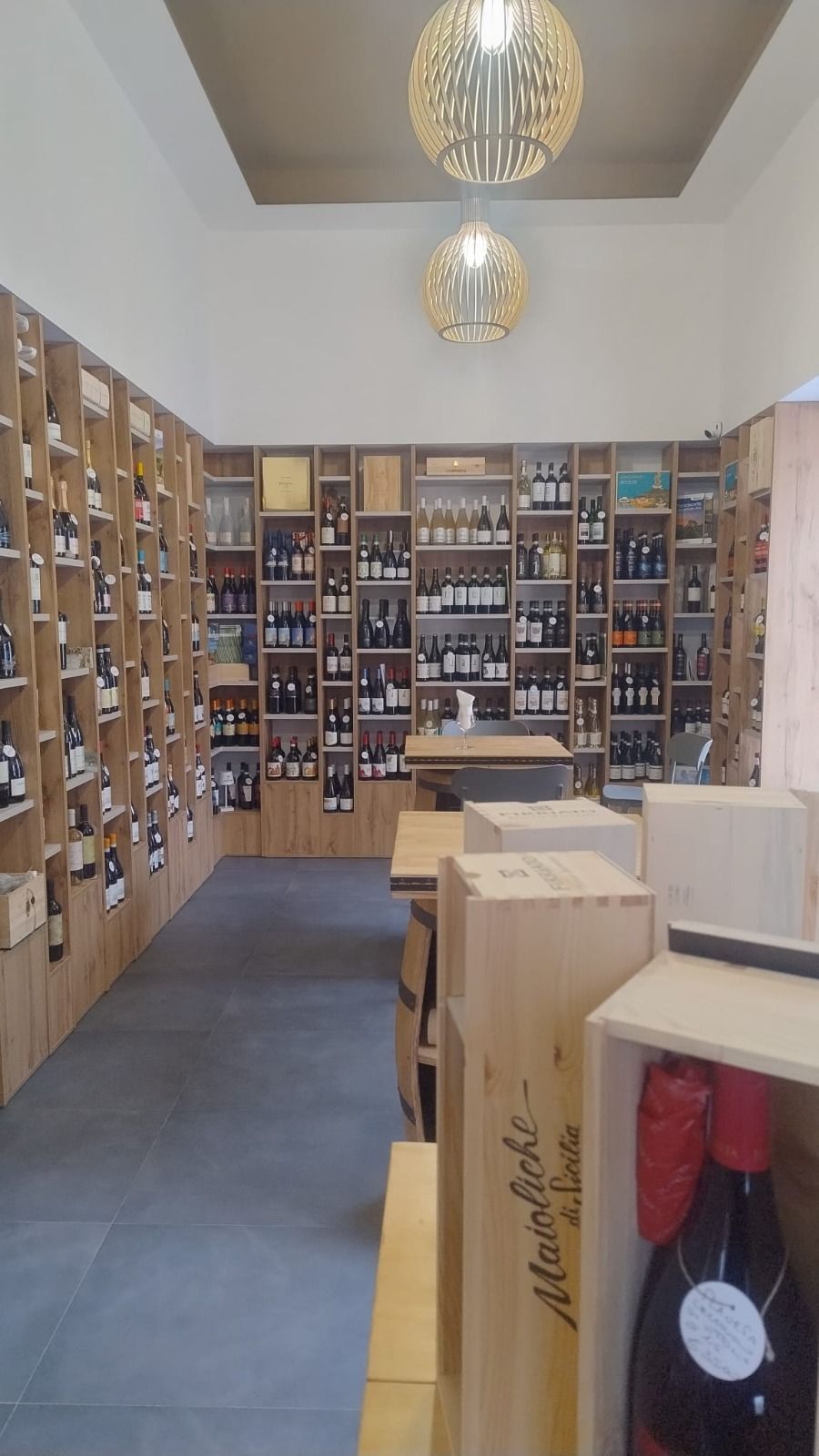 L'interno di un'enoteca è caratterizzato da scaffalature in legno a tutta altezza piene di bottiglie di vino, due lampade a globo e casse.