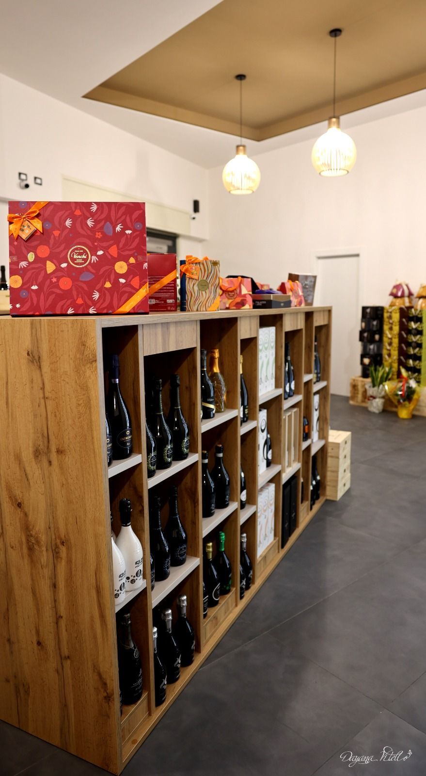 Una scaffalatura in legno piena di bottiglie di vino in un negozio, con due lampade a sospensione sopra.