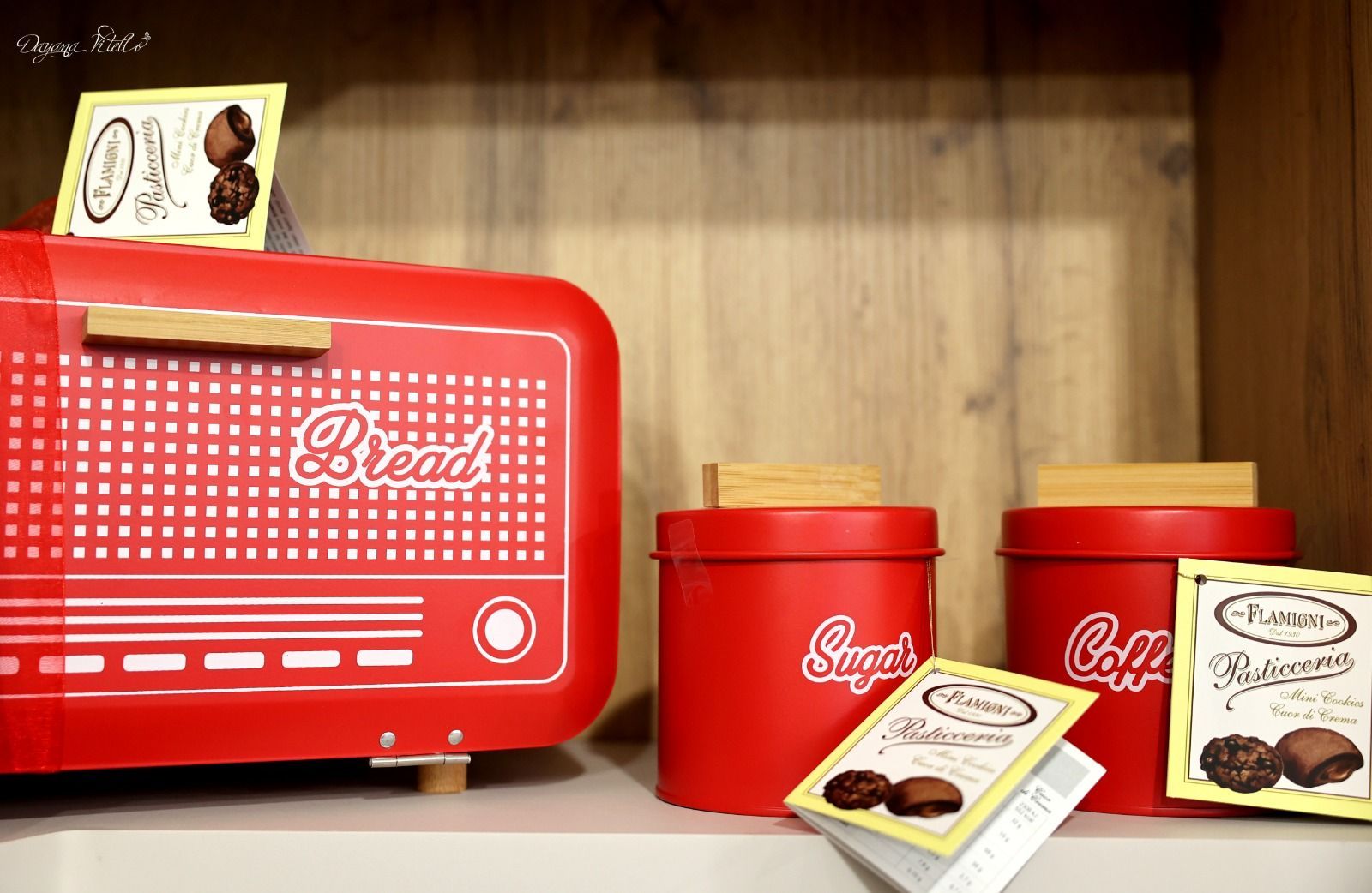 Su uno scaffale di legno si trovano un portapane rosso acceso e dei barattoli coordinati per zucchero e caffè, con manici in legno chiaro.
