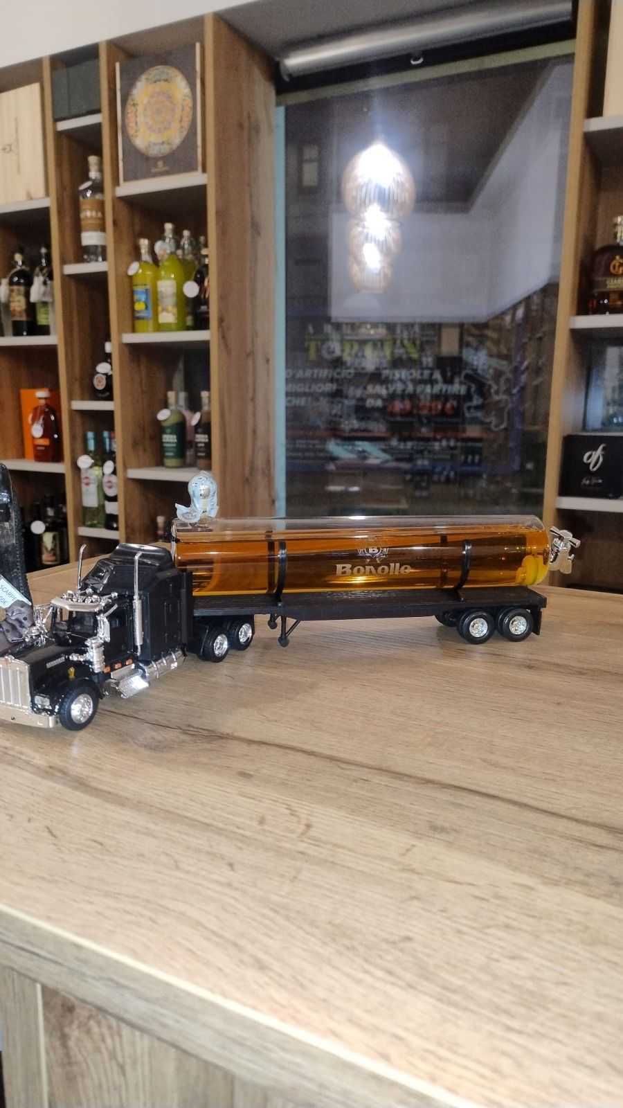 Un modellino di camion con un liquido color ambra è appoggiato su un bancone di legno chiaro davanti a uno scaffale di liquori.