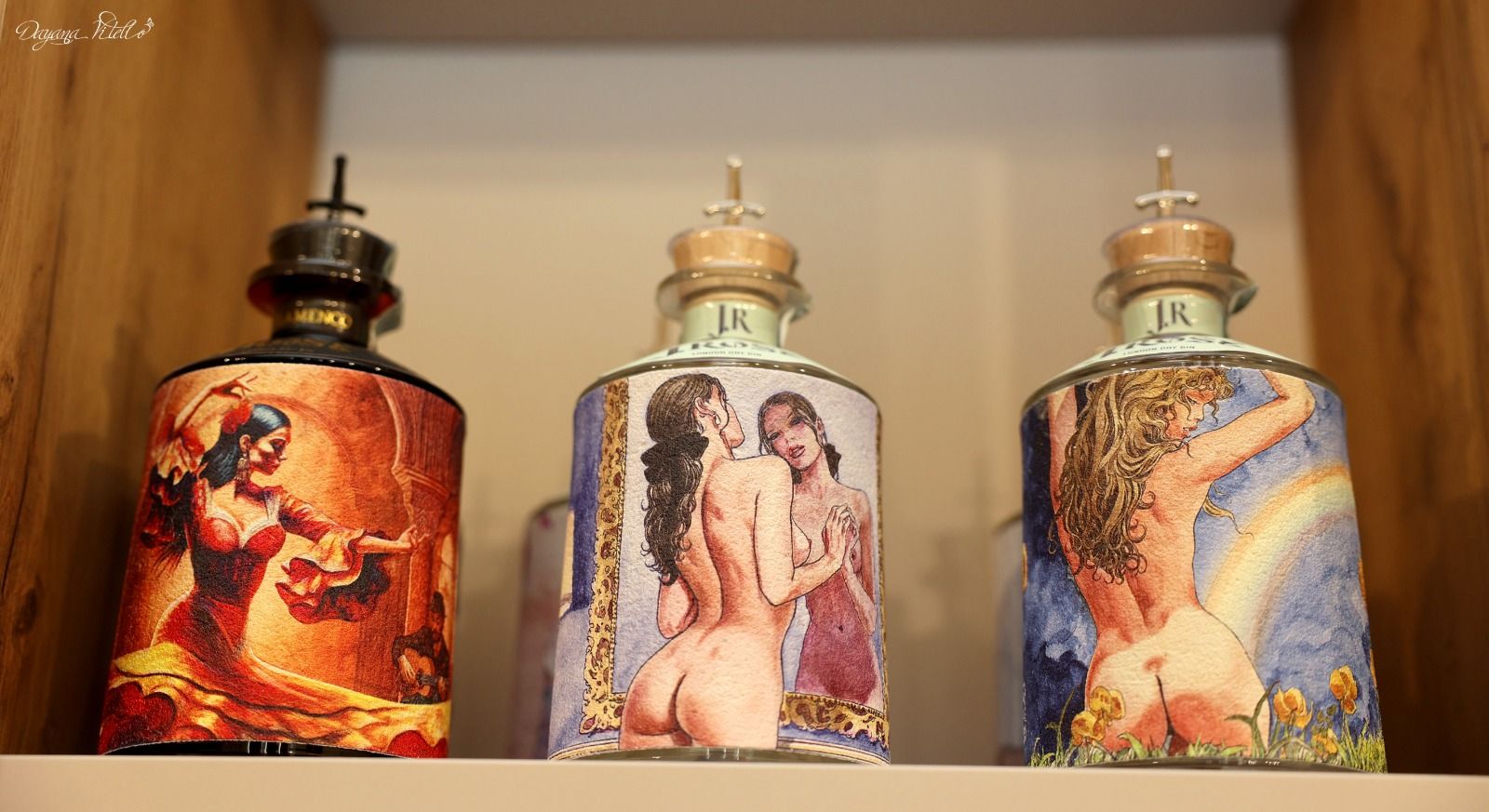 Tre bottiglie cilindriche con etichette decorative raffiguranti illustrazioni, disposte su uno scaffale di legno.