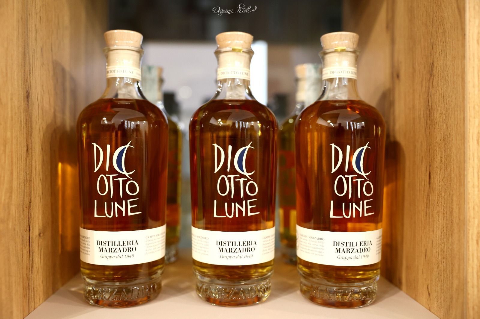 Tre bottiglie di grappa Diciotto Lune dal colore ambrato e con tappo di sughero, esposte su uno scaffale di legno.