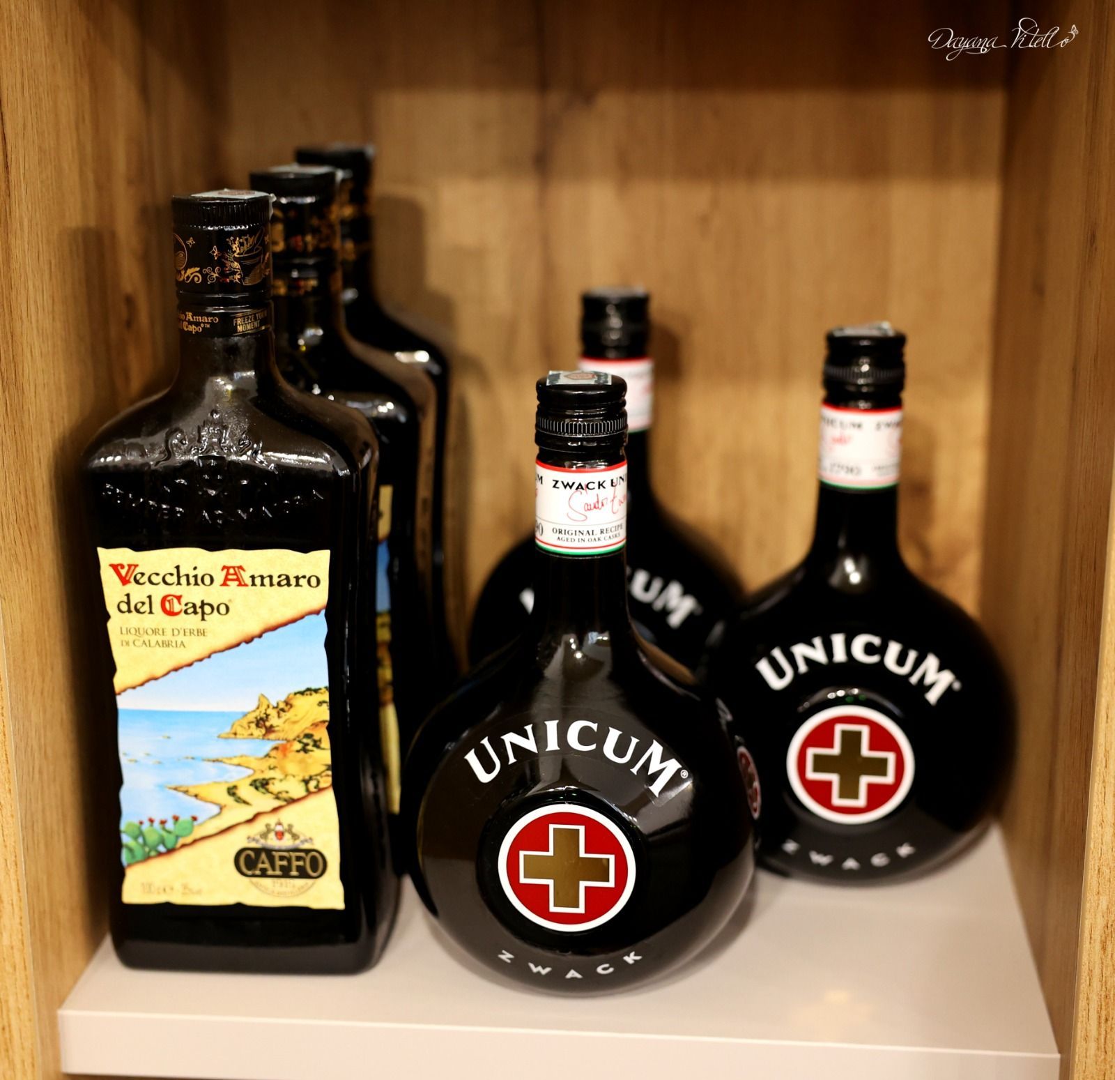 Una collezione di bottiglie di liquore Unicum e Vecchio Amaro del Capo disposte su uno scaffale di legno.