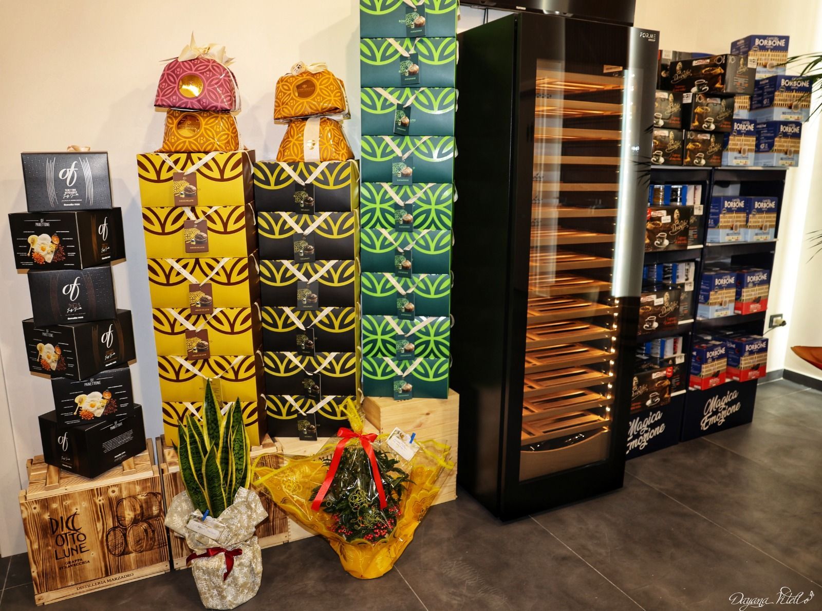 All'interno di un negozio, scatole regalo impilate nei colori giallo, nero e verde sono disposte accanto a un frigorifero alto e a degli scaffali.