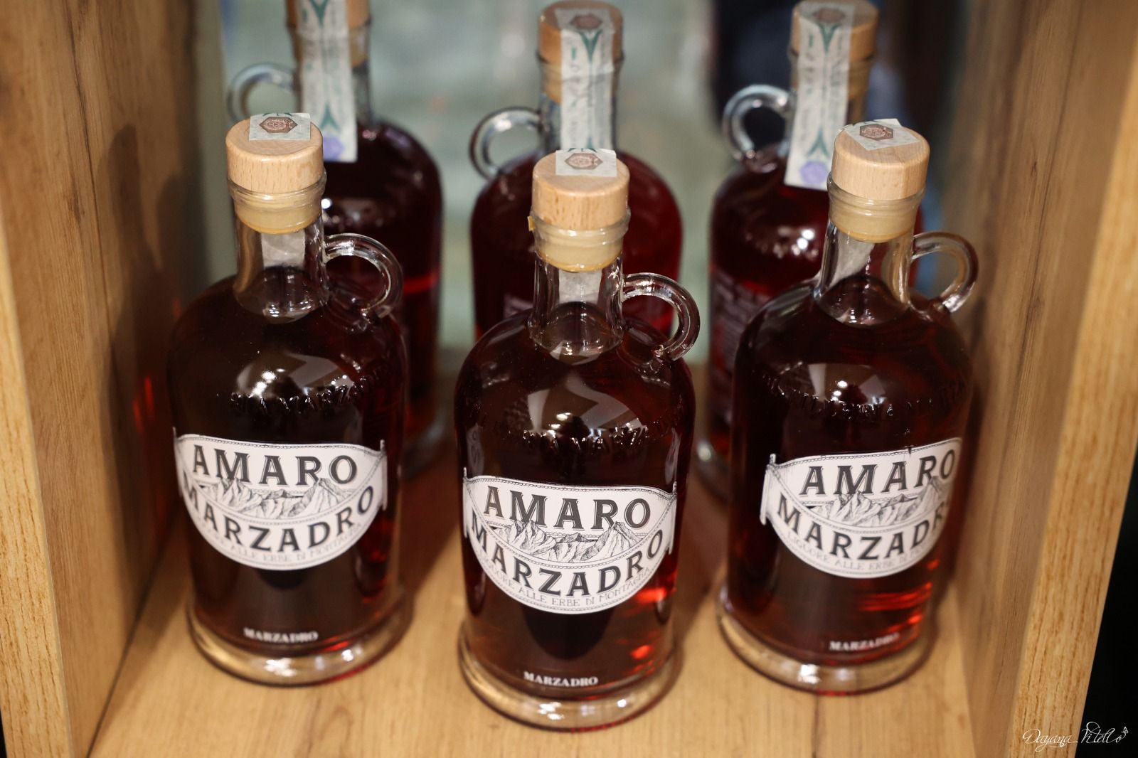 Cinque bottiglie di liquore alle erbe Amaro Marzadro, disposte su scaffali di legno, con tappi di sughero e liquido ambrato.