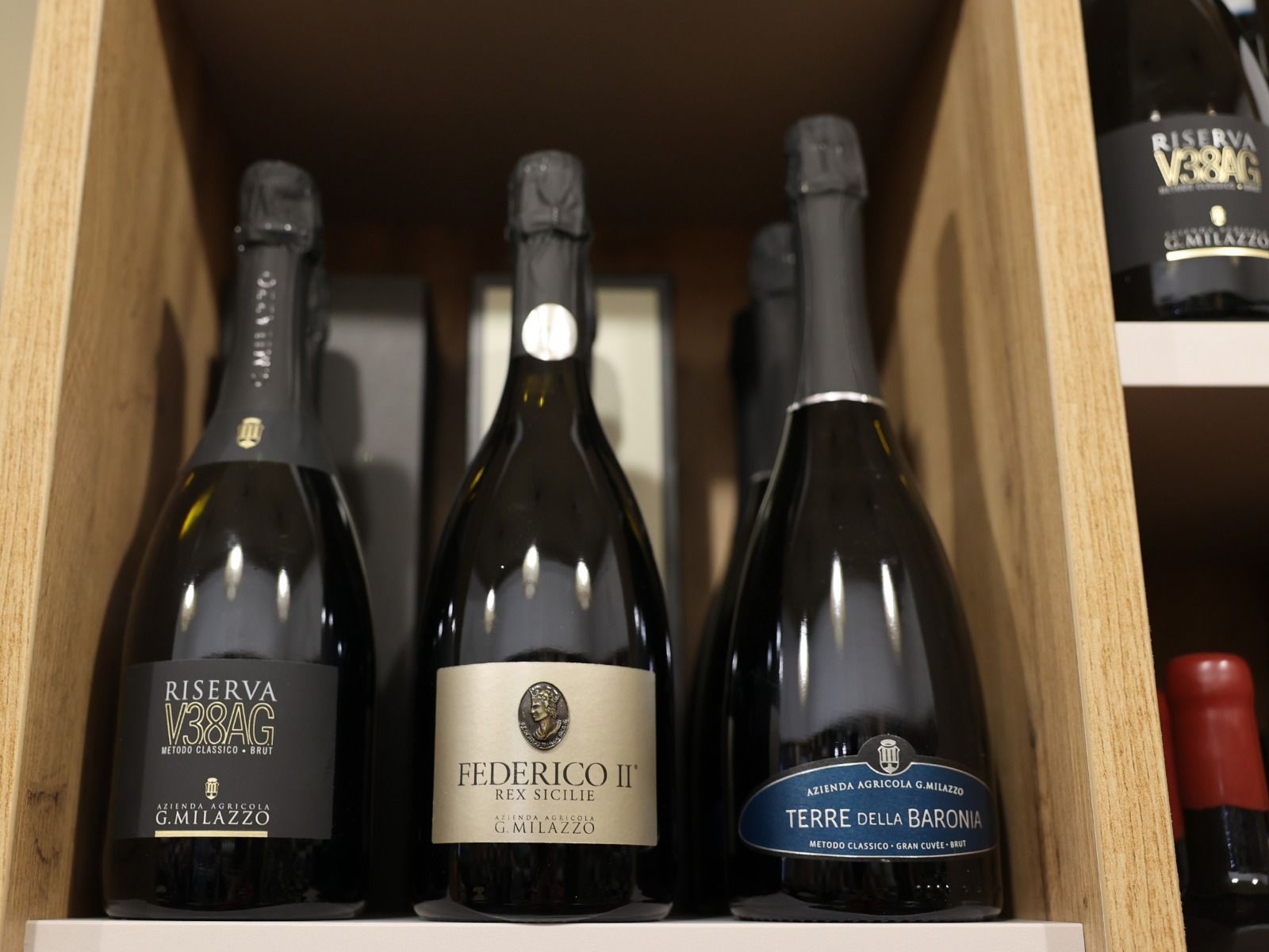 In un negozio, tre bottiglie di spumante con etichette diverse sono appoggiate su uno scaffale di legno.