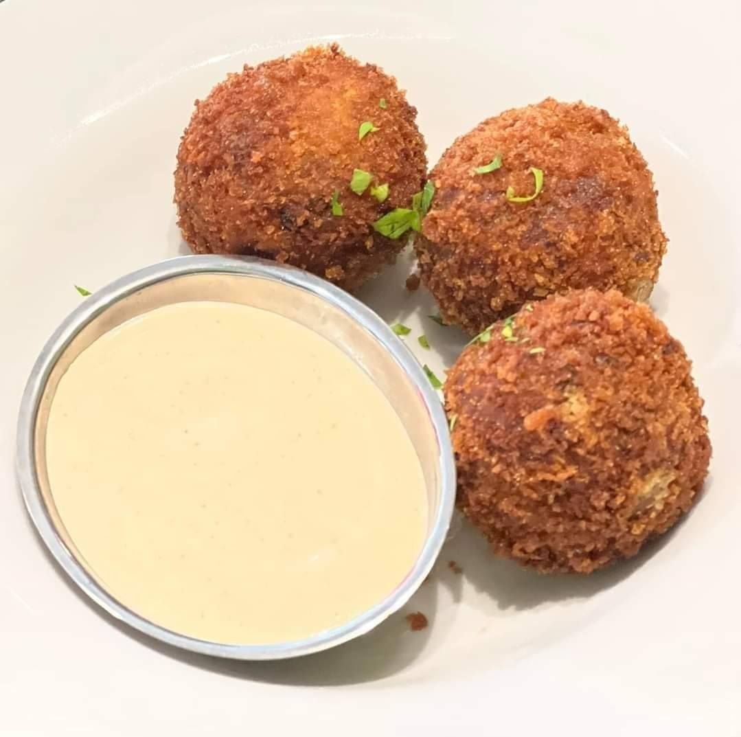 Three golden-brown fried balls on a white plate, served with a small bowl of creamy sauce.